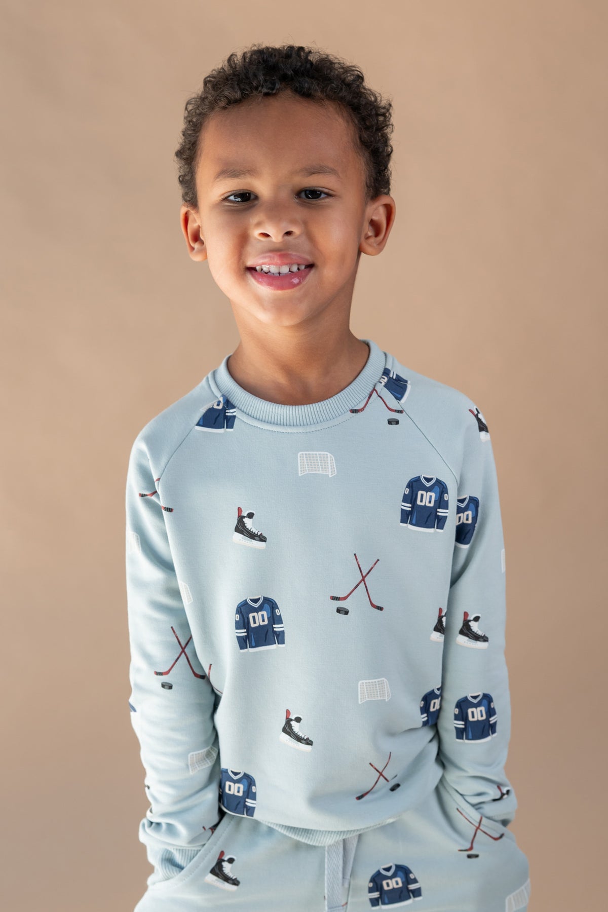 A young boy smiling, wearing a light blue long-sleeve sweatshirt and matching pants with an all-over hockey print of blue jerseys, skates, and sticks, set against a solid beige background.