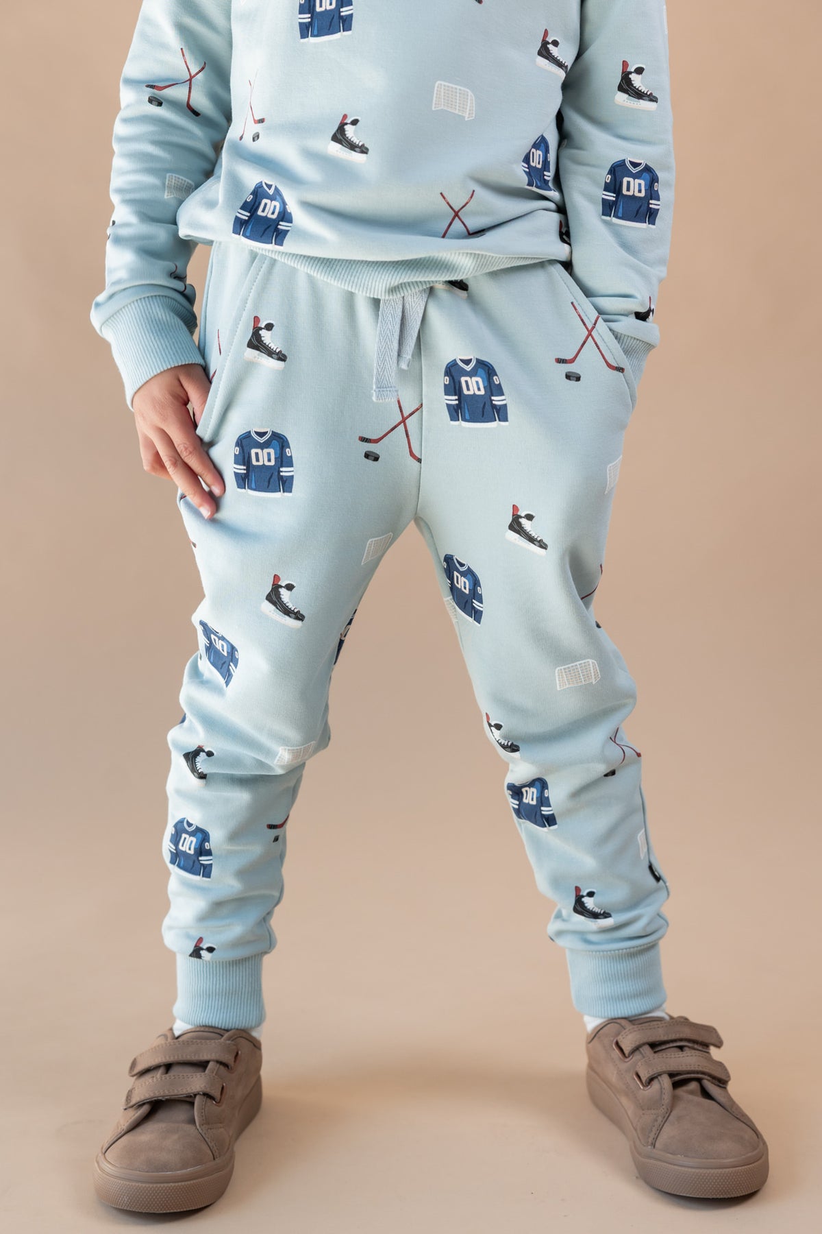A child wearing a light blue matching sweatshirt and jogger pants set with a hockey-themed print of jerseys, skates, and sticks, paired with brown suede sneakers, against a solid beige background.
