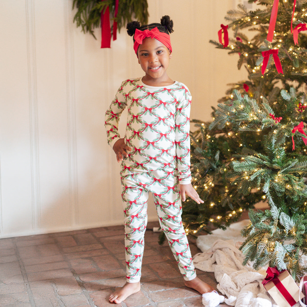 Young girl standing beside a decorated Christmas tree wearing the Long Sleeve Pajamas in Holiday Bow with a red knotted bow headband