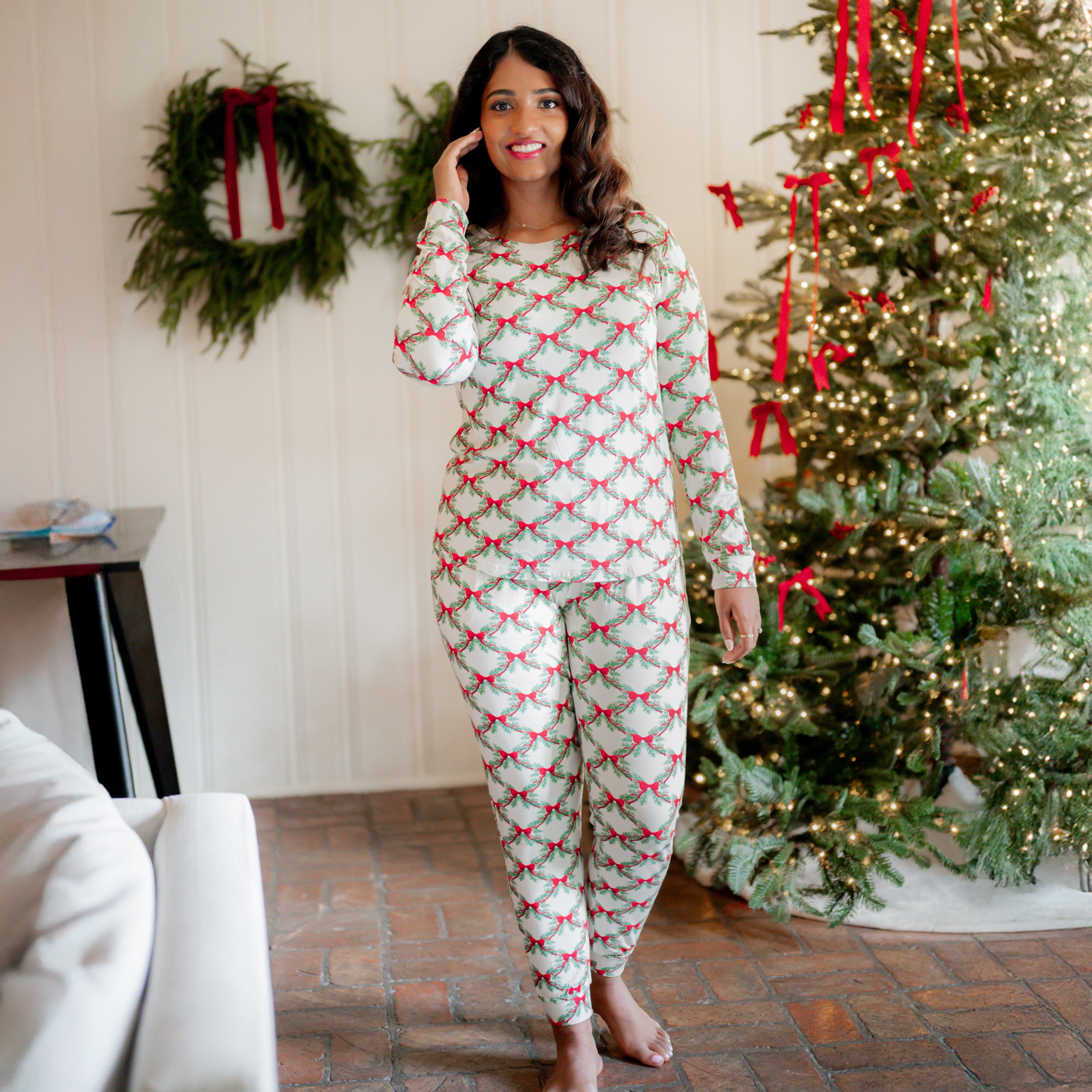 Female model standing in front of a white paneled wall and decorated Christmas tree wearing the Women's Jogger Pajama Set in Holiday Bow