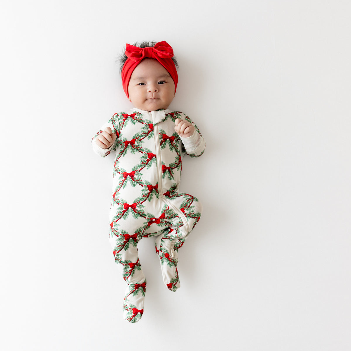 Infant laying down wearing the Zippered Footie in Holiday Bow with a Bow Headband in Cardinal