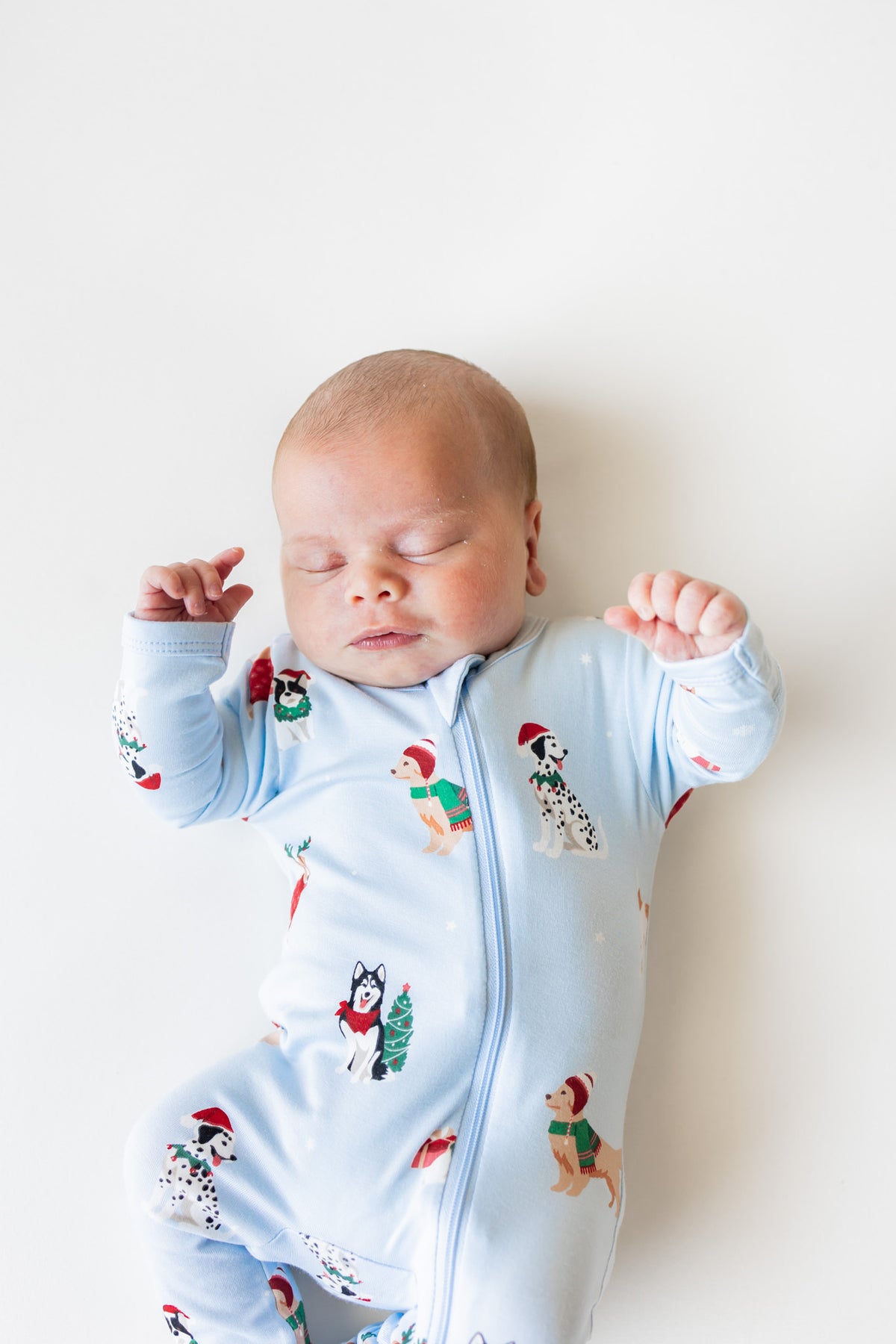 A sleeping newborn baby wearing a light blue zip-up footed sleeper with a festive print of Dalmatian dogs in Christmas outfits, lying on a plain white background.