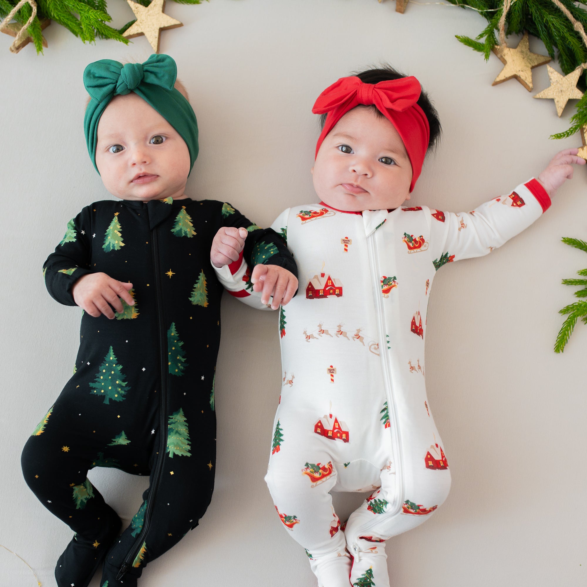 Two infants laying side by side with their arms intertwined wearing zippered footies in Santa Sleigh and twinkle trees with Bow Headband in Cardinal and Emerald
