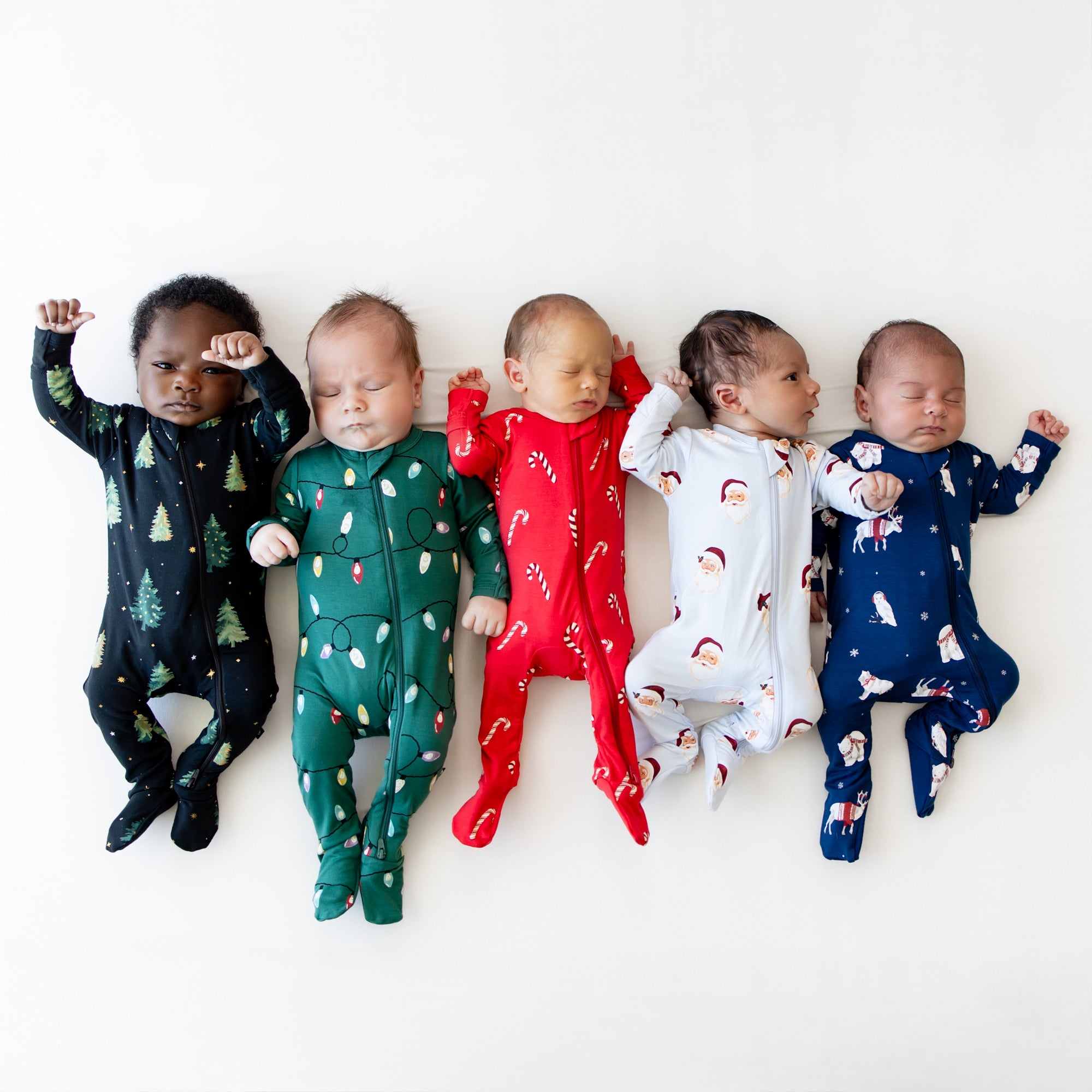 Five infant models laying side by side wearing zippered footies in Zippered Footie in Frosty Friends, Vintage Santa, Candy Cane, Merry and Bright, and Twinkle Trees