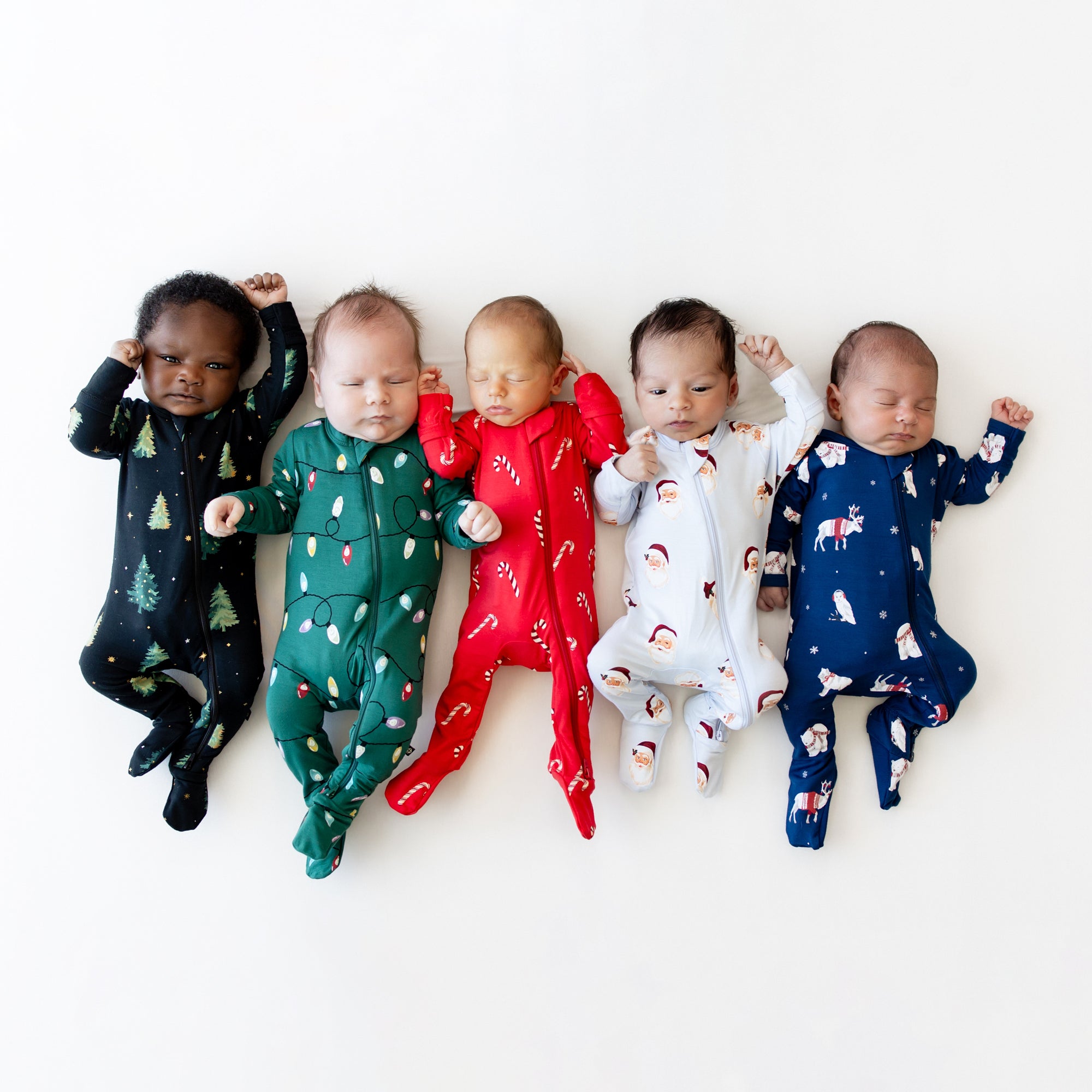 Five infant models laying side by side wearing Zippered Footie in Candy Cane, Vintage Santa, Frosty Friends, Merry and Bright and Twinkle Tree