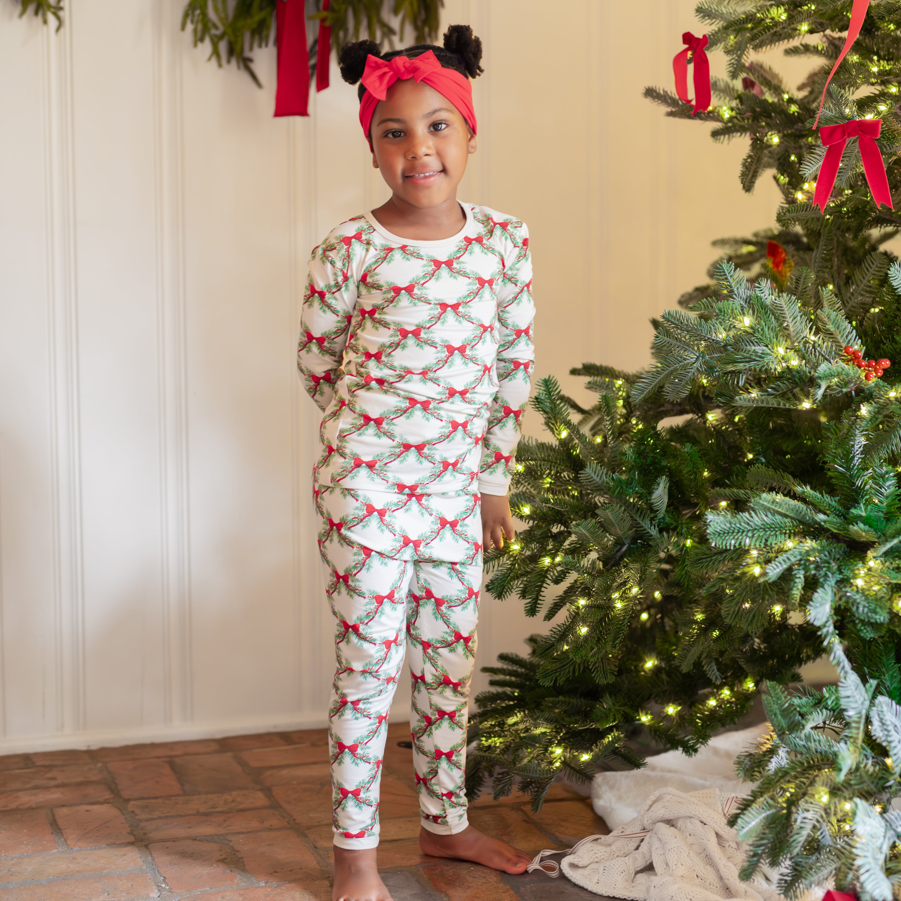 Young girl standing beside a decorated Christmas tree wearing the Long Sleeve Pajamas in Holiday Bow with a Cardinal knotted bow headband