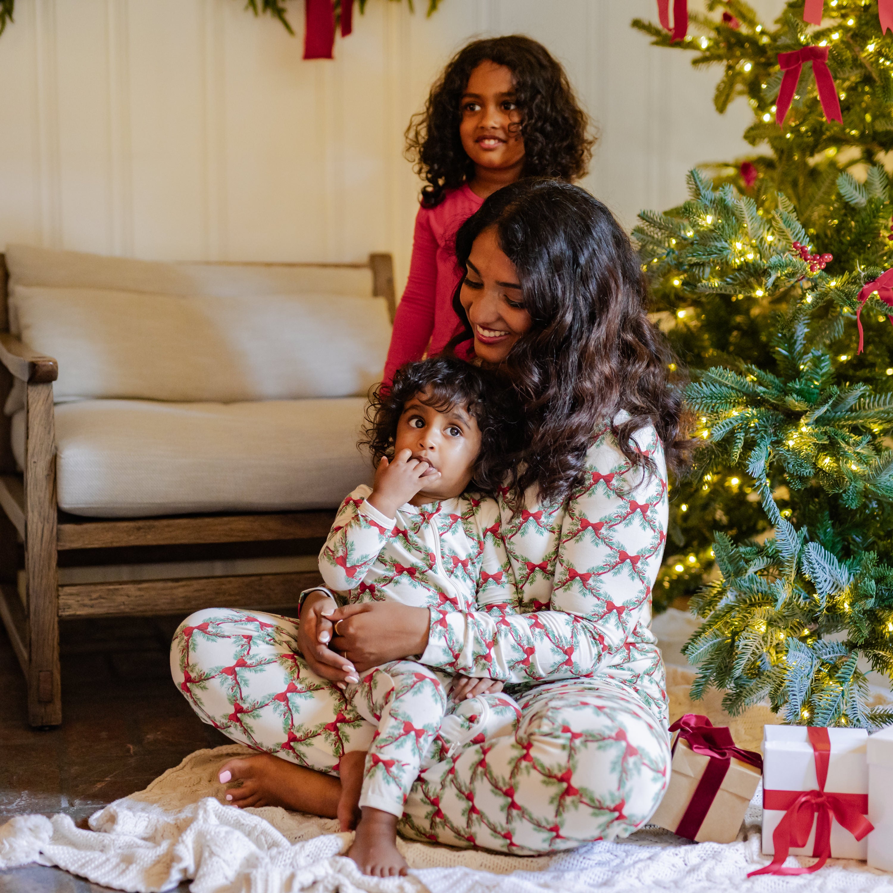 Mother wearing the Women's Jogger Pajama Set in Holiday Bow sitting on the floor beside a decorated Christmas tree holding her toddler who is wearing the matching zippered romper in front of older child wearing the long sleeve toddler pajama set in cardinal
