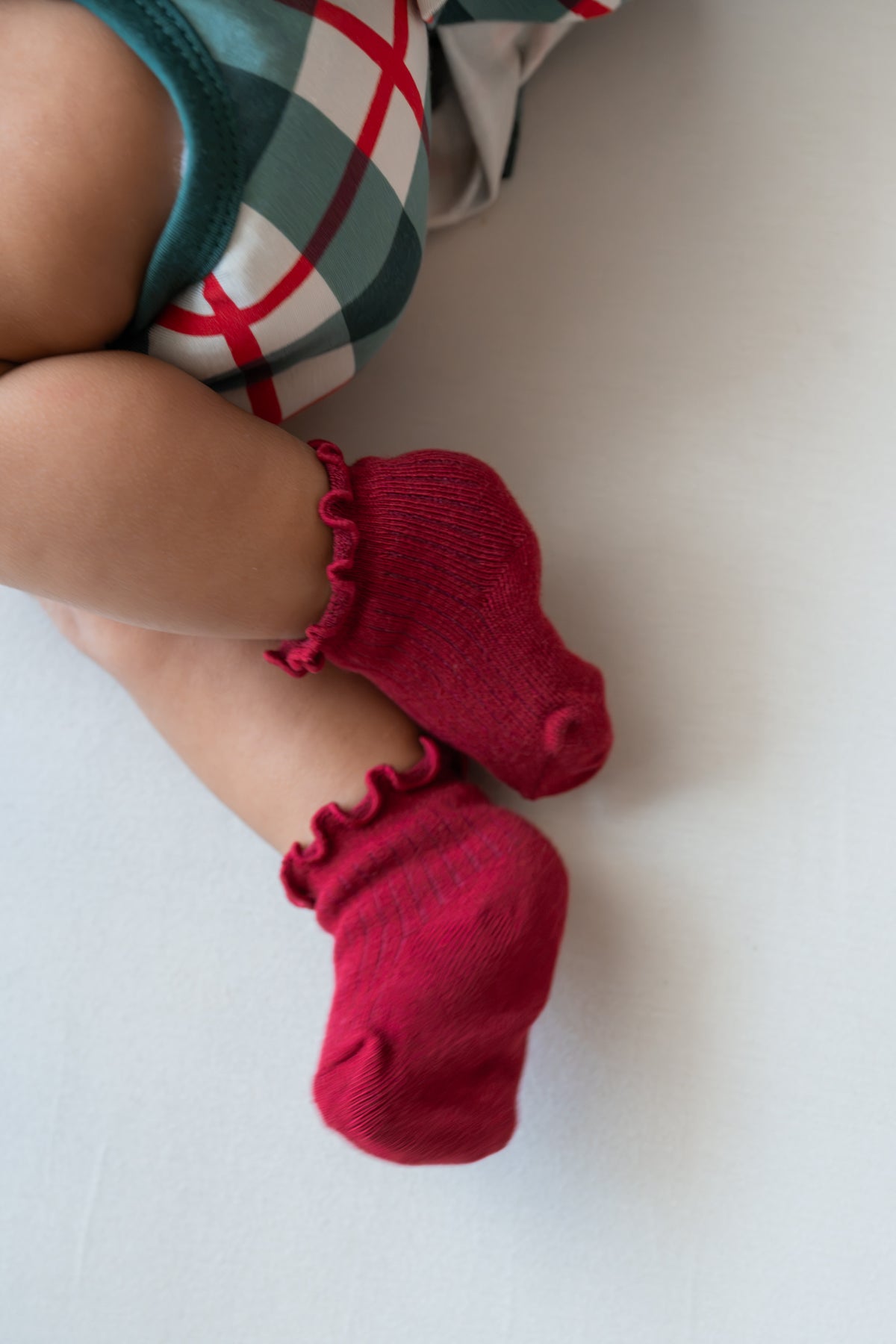 A close-up of a baby's legs wearing bright red ruffled ankle socks, with a plaid bodysuit partially visible.