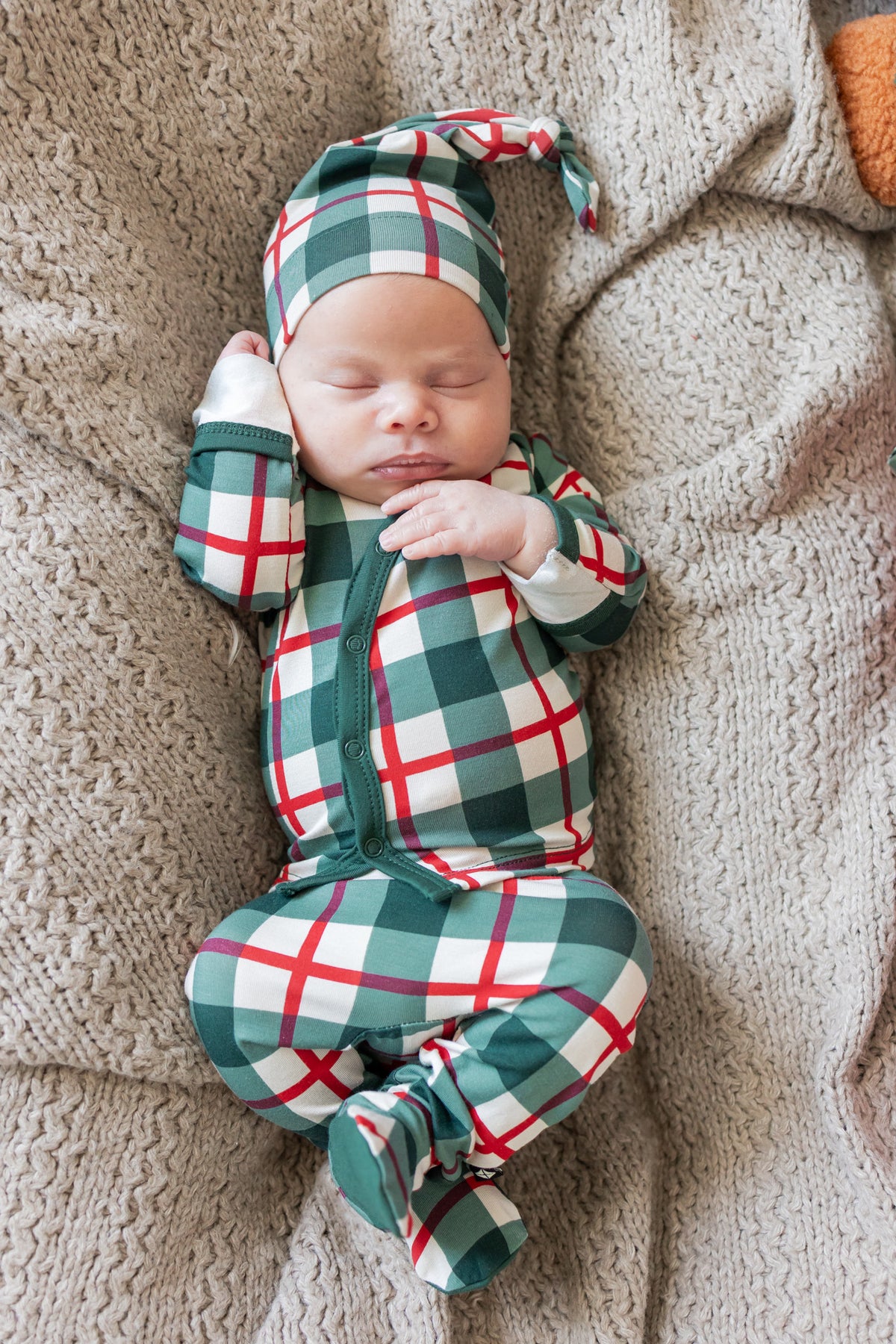 A sleeping newborn baby wearing a green, red, and white plaid footed set and matching pointed hat, lying on a cozy beige knit blanket.