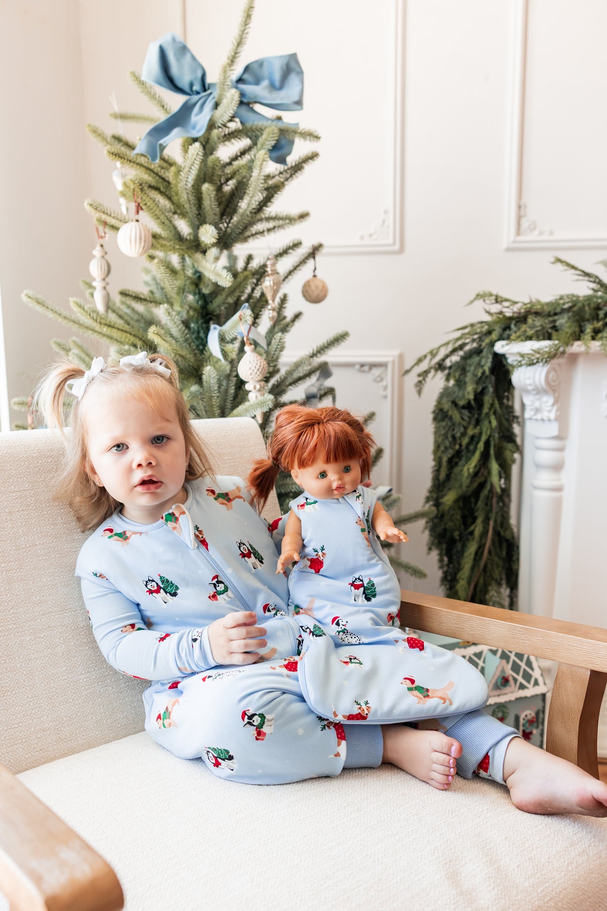 Girl sitting on sofa wearing Sleep Bag Walker in Holiday Pups 1.0 print holding a doll wearing a similar print baby doll sleep bag