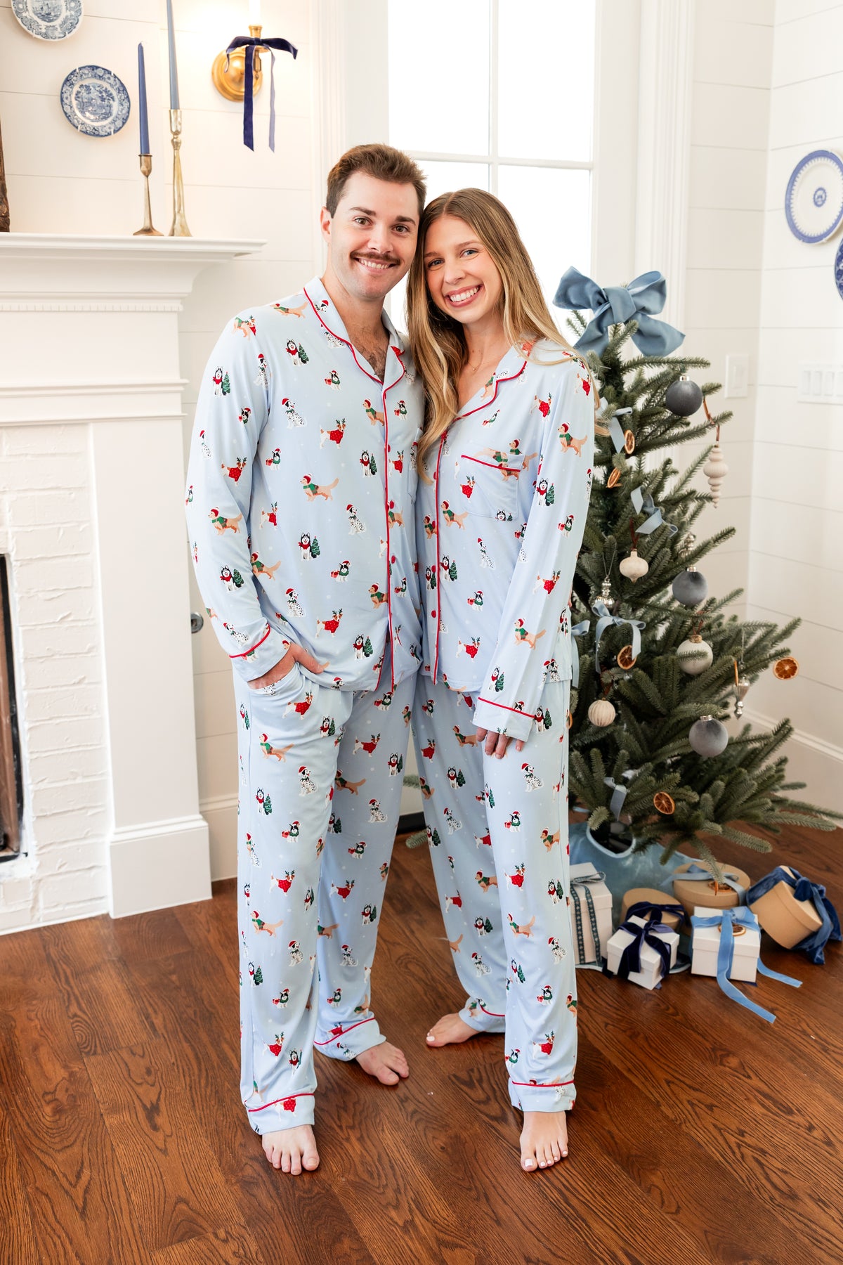 A man and a woman standing barefoot in a living room, wearing matching light blue pajama sets with a festive Christmas print and red piping. A decorated Christmas tree, fireplace, and wrapped gifts are visible in the background.
