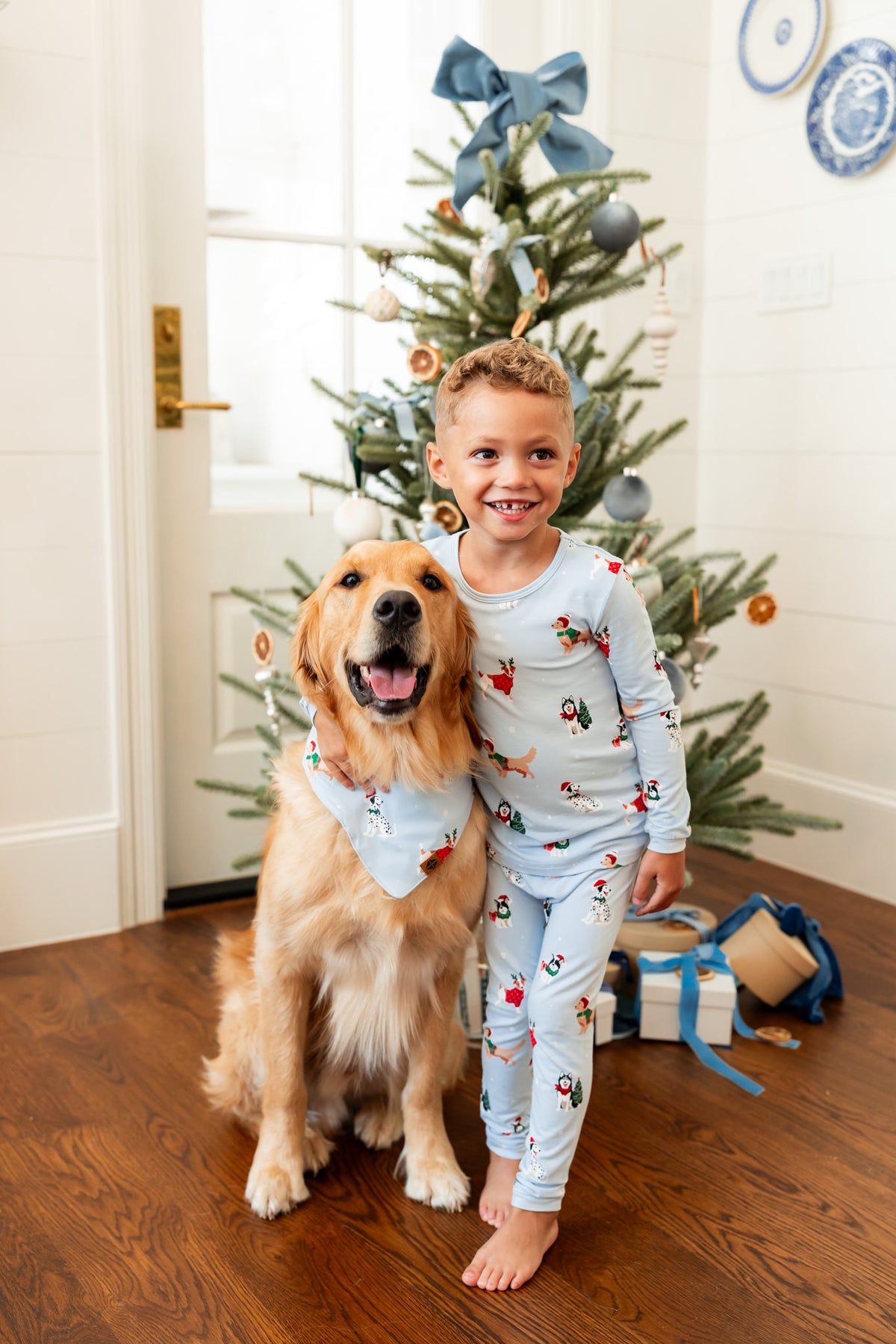 Boy wearing Long Sleeve Pajamas in Holiday Pups print while hugging the dog beside him wearing similar print dog bandana, with Christmas tree and presents in the background