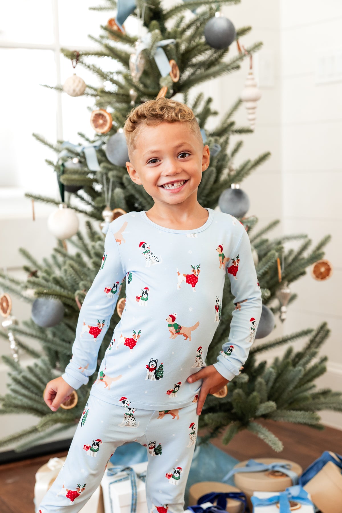 Boy posing in Long Sleeve Pajamas in Holiday Pups print with Christmas tree and presents in the background