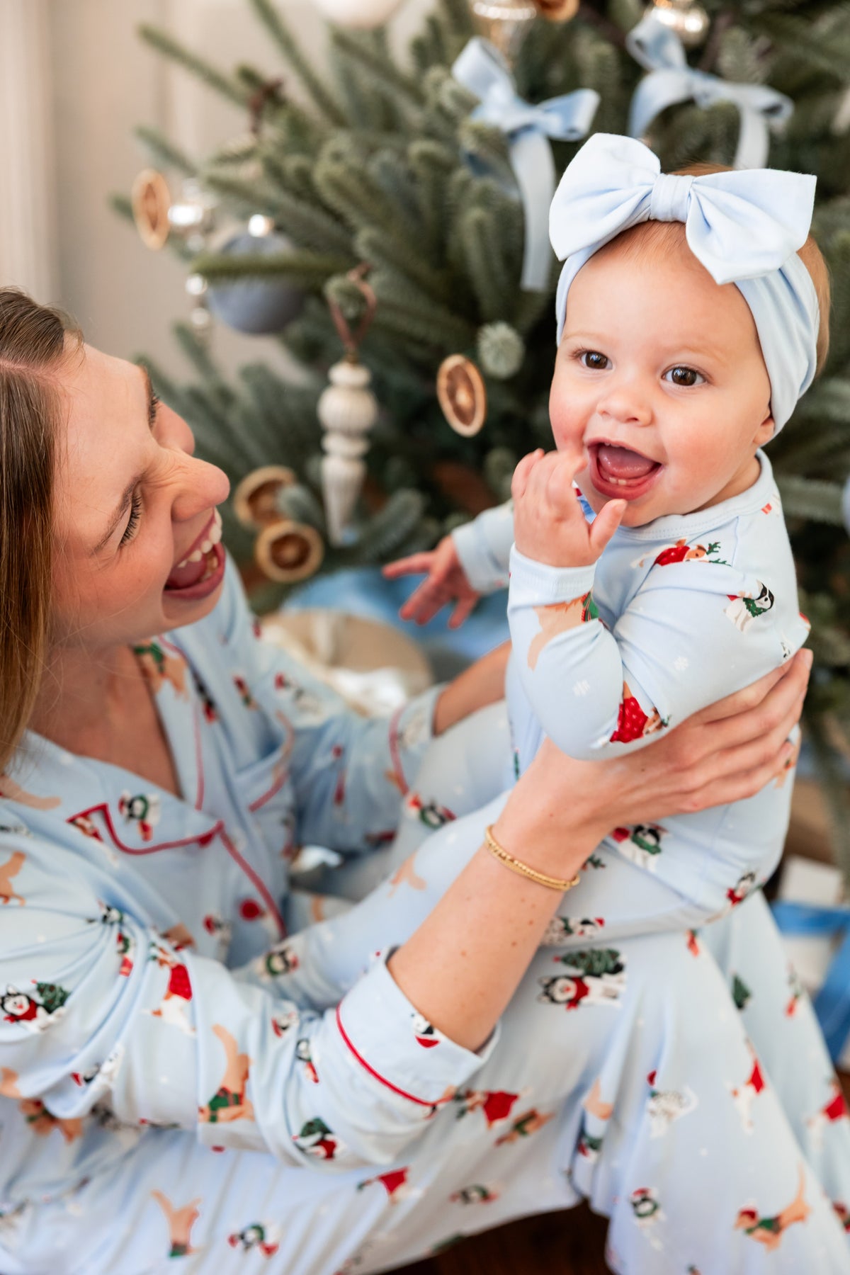 A woman and a baby wearing matching light blue Christmas-themed pajamas sit together in front of a decorated Christmas tree. The pajamas have a festive print with red accents, and the baby also wears a matching light blue bow headband.