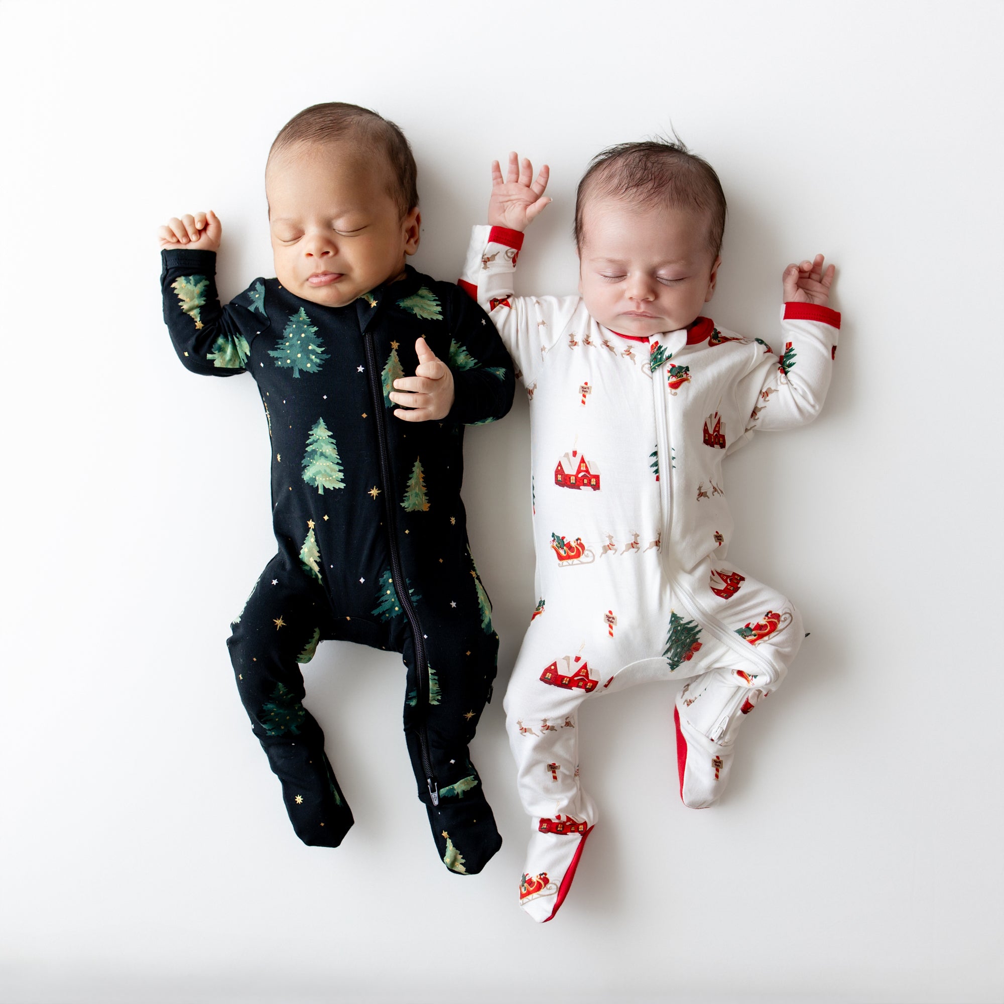 Two newborns laying side by side wearing the Zippered Footie in Santa Sleigh and twinkle trees