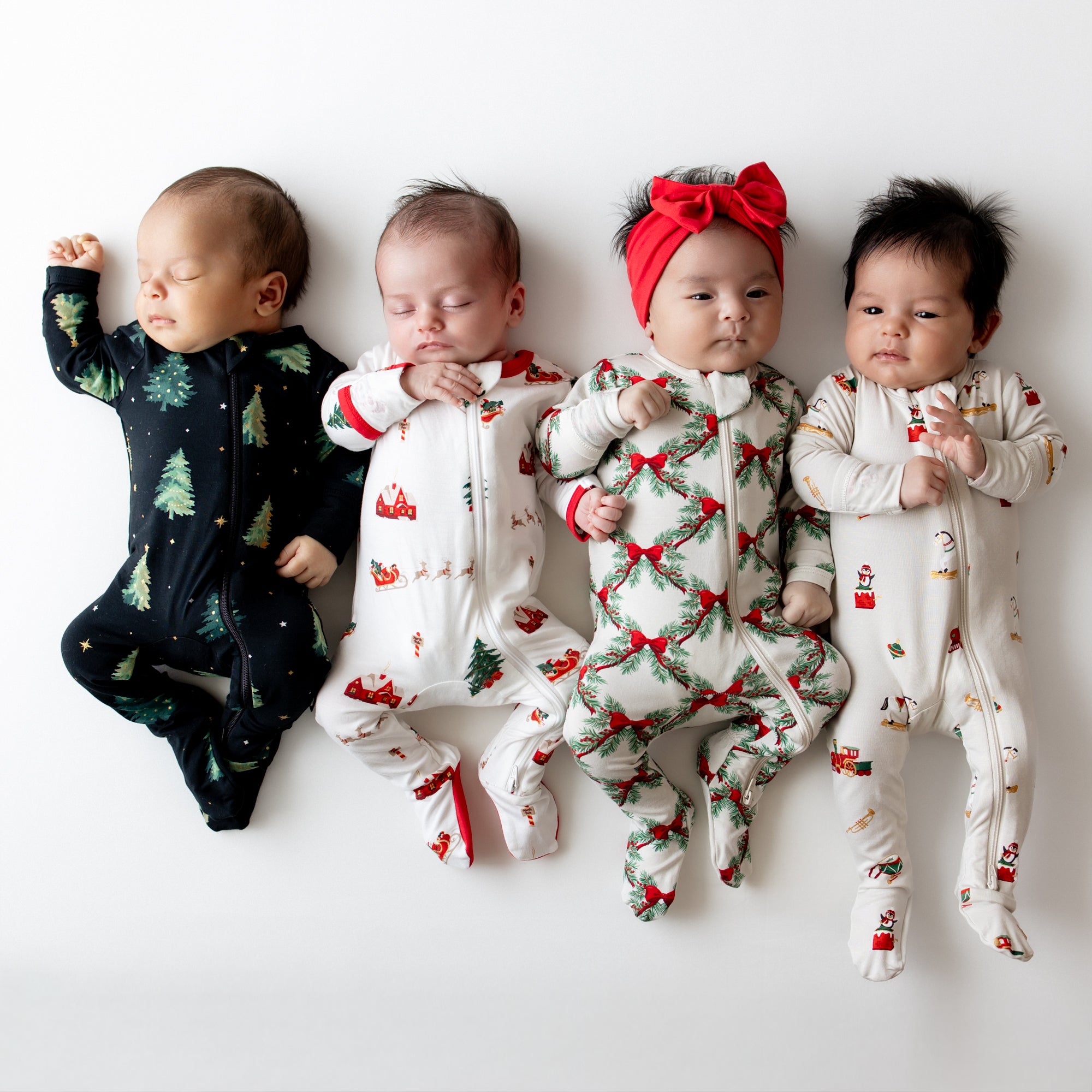 Four infants laying side by side on a white blanket wearing the Zippered Footie in Holiday Bow, Vintage Toys, Santa Sleigh and Twinkle Trees