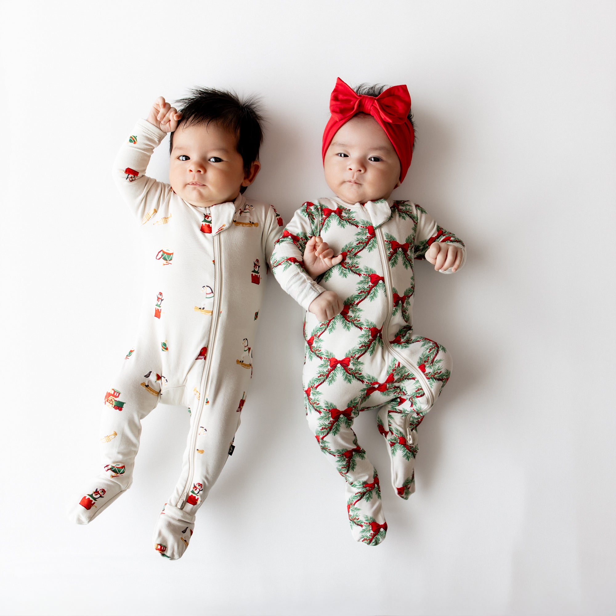 Two infants laying size by side wearing the Zippered Footie in Holiday Bow and Vintage Toys