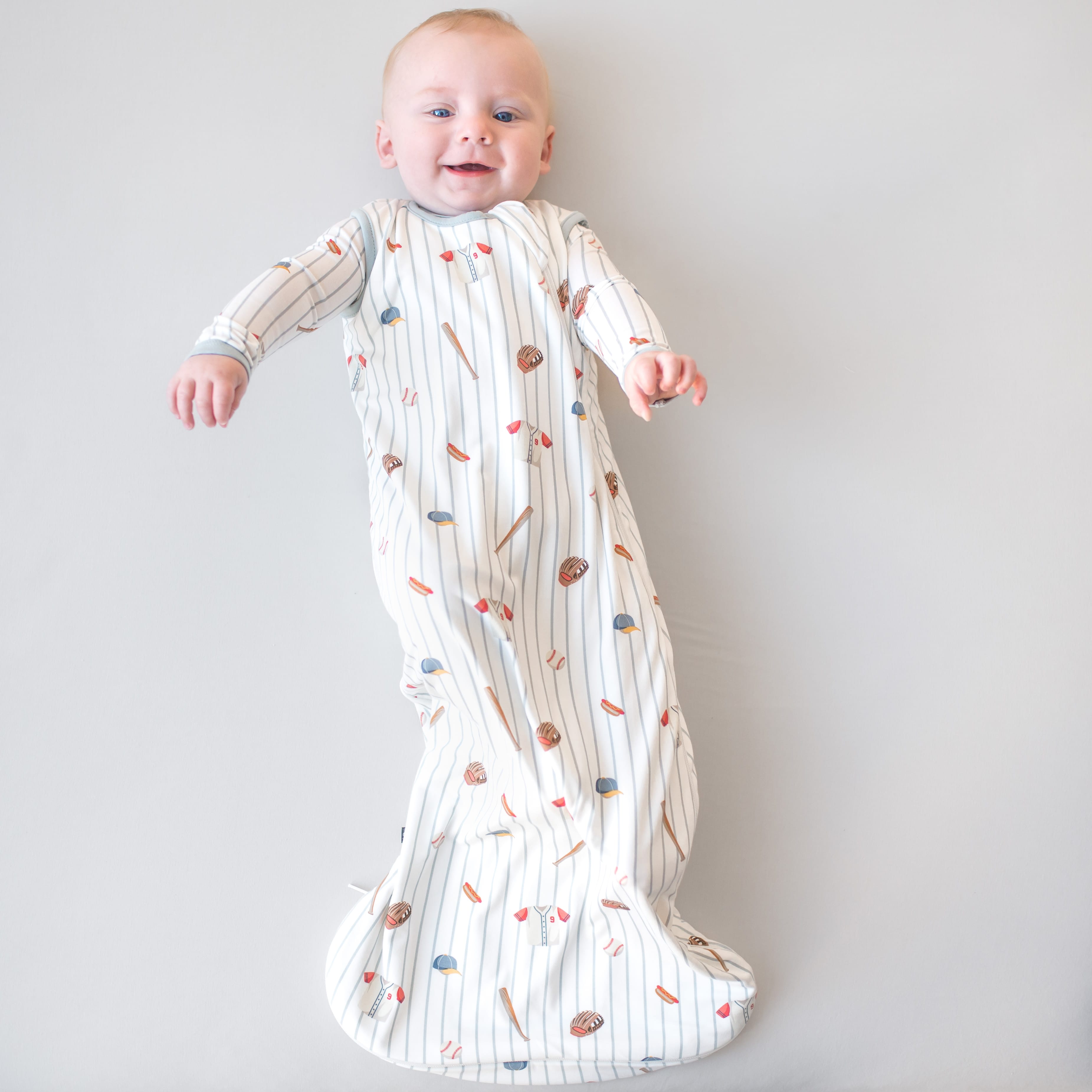 Infant laying on a light neutral surface wearing the Sleep Bag in Home Run 0.5 with matching romper on underneath