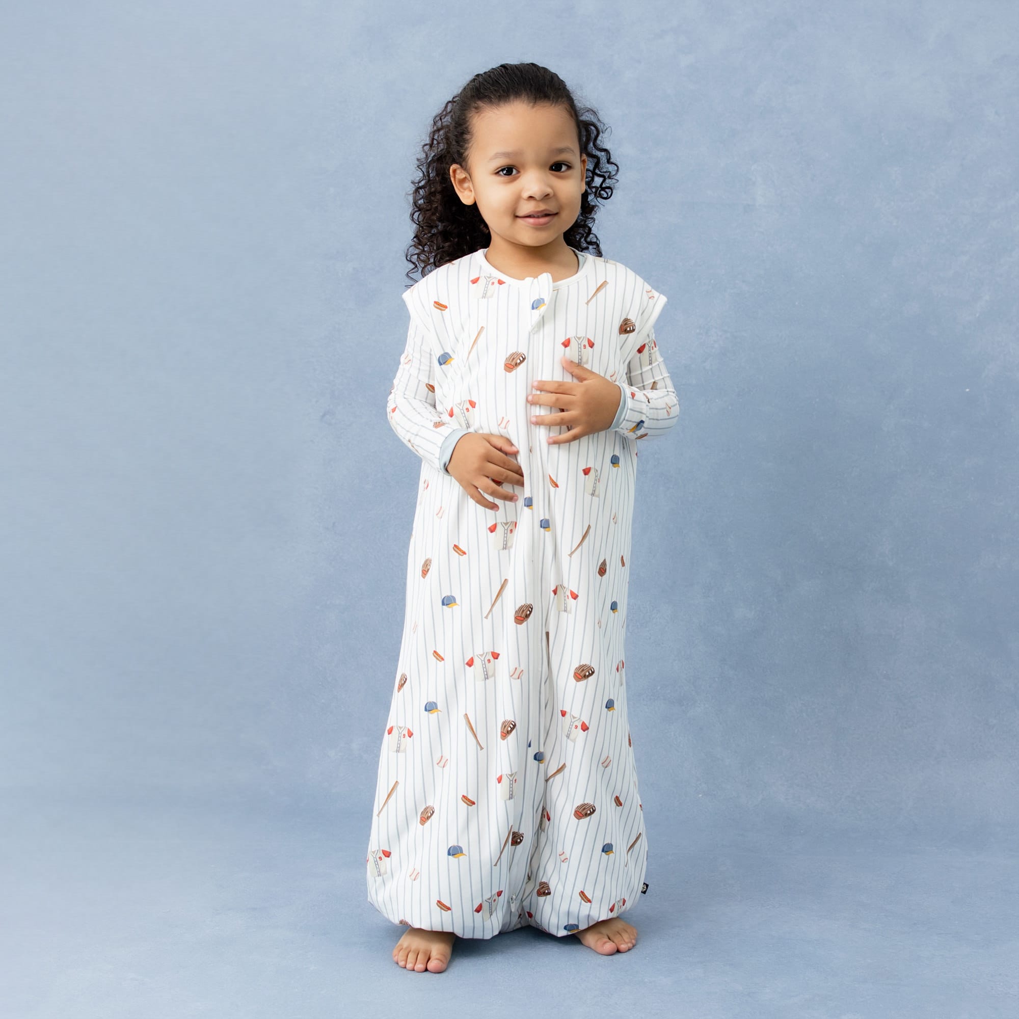 Young girl wearing the Sleep Bag Walker in Home Run 1.0 with matching long sleeve pajamas on underneath standing in front of a marbled blue background
