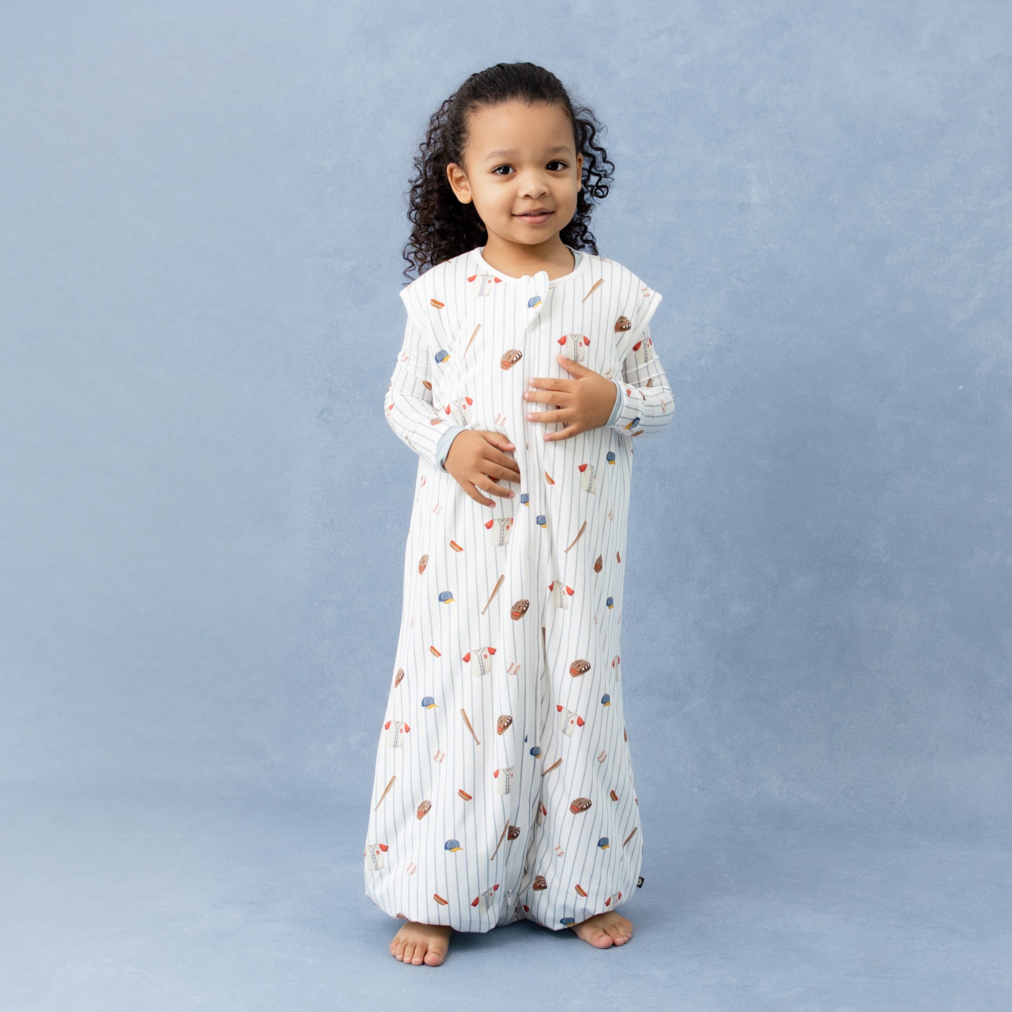 Young girl wearing the Sleep Bag Walker in Home Run 1.0 with matching long sleeve pajamas on underneath standing in front of a marbled blue background