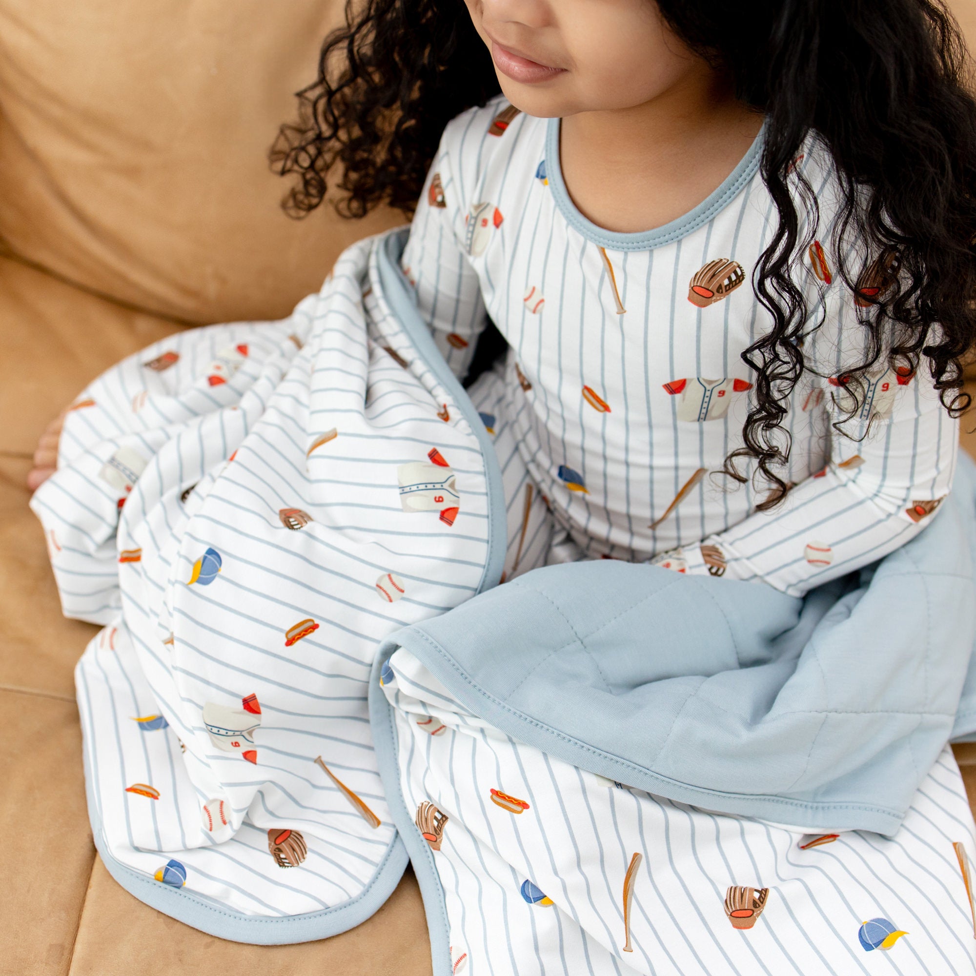 Close up of the Toddler Blanket in Home Run 1.0 around a girl sitting on a brown couch wearing matching long sleeve pajamas