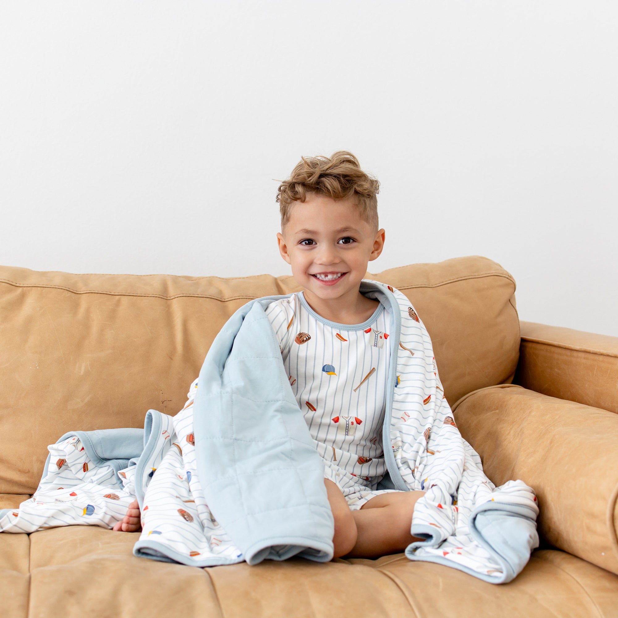 Young boy sitting on a brown could with the Youth Blanket in Home Run 1.0 wrapped around his shoulder wearing the matching short sleeve pajama set