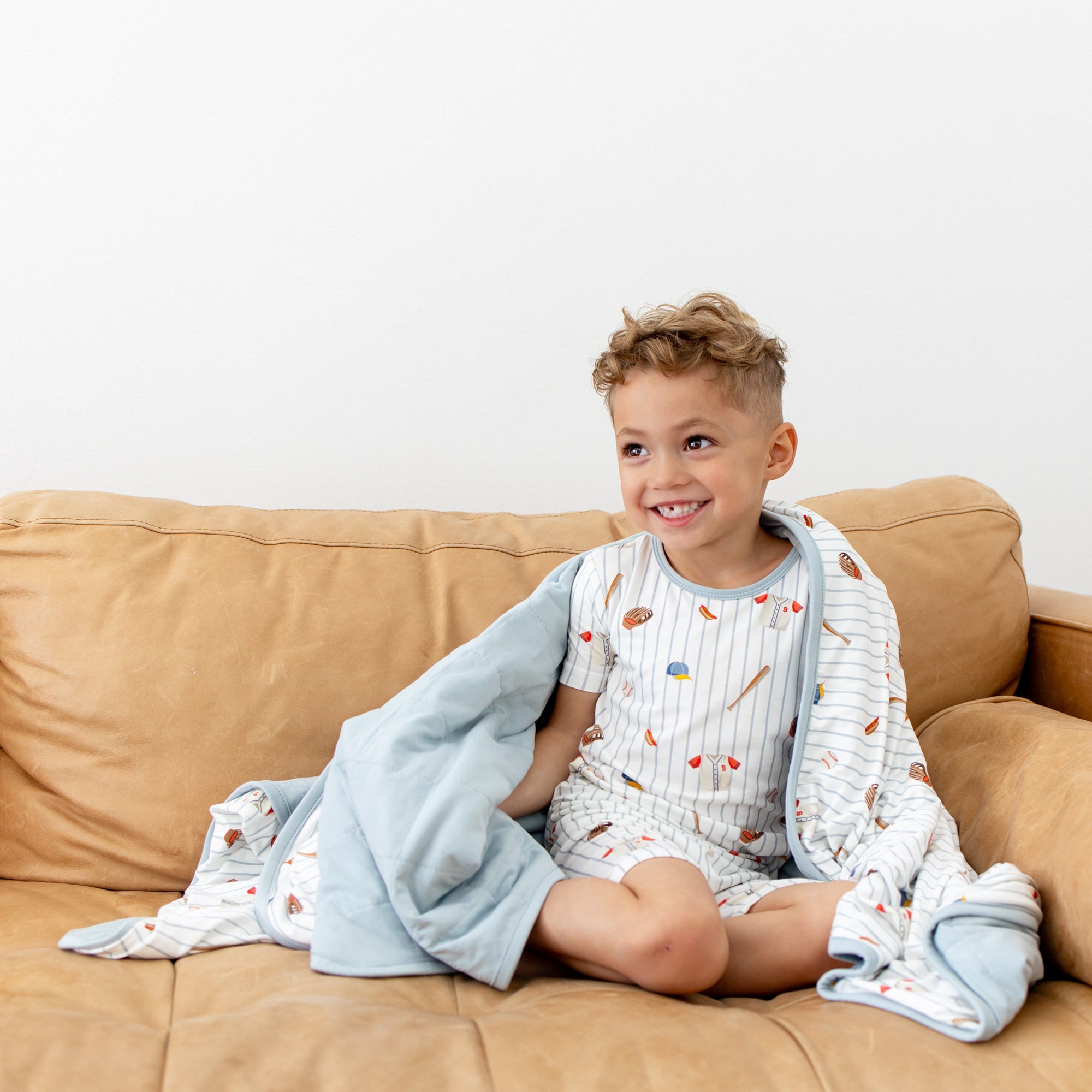 Young boy sitting on a brown couch with the Youth Blanket in Home Run 1.0 around him wearing matching short sleeve pajamas
