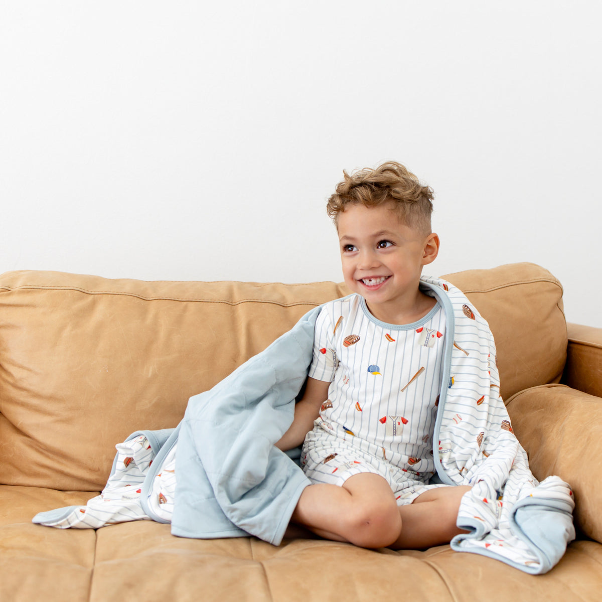Young boy sitting on a brown couch with the Youth Blanket in Home Run 1.0 around him wearing matching short sleeve pajamas