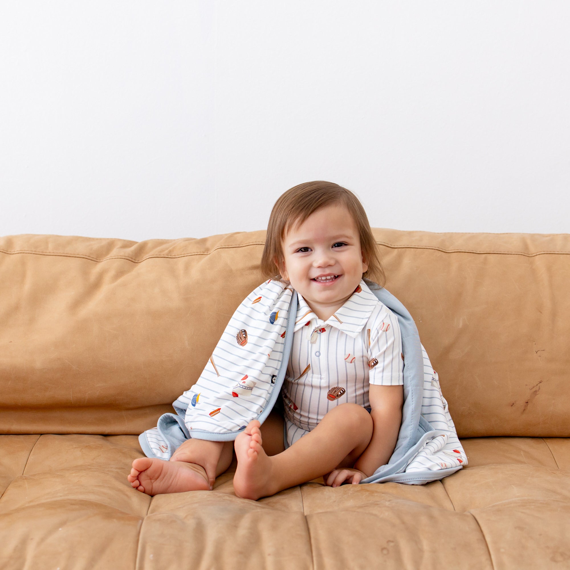 Young boy sitting on a brown couch with the Baby Blanket in Home Run around his shoulders