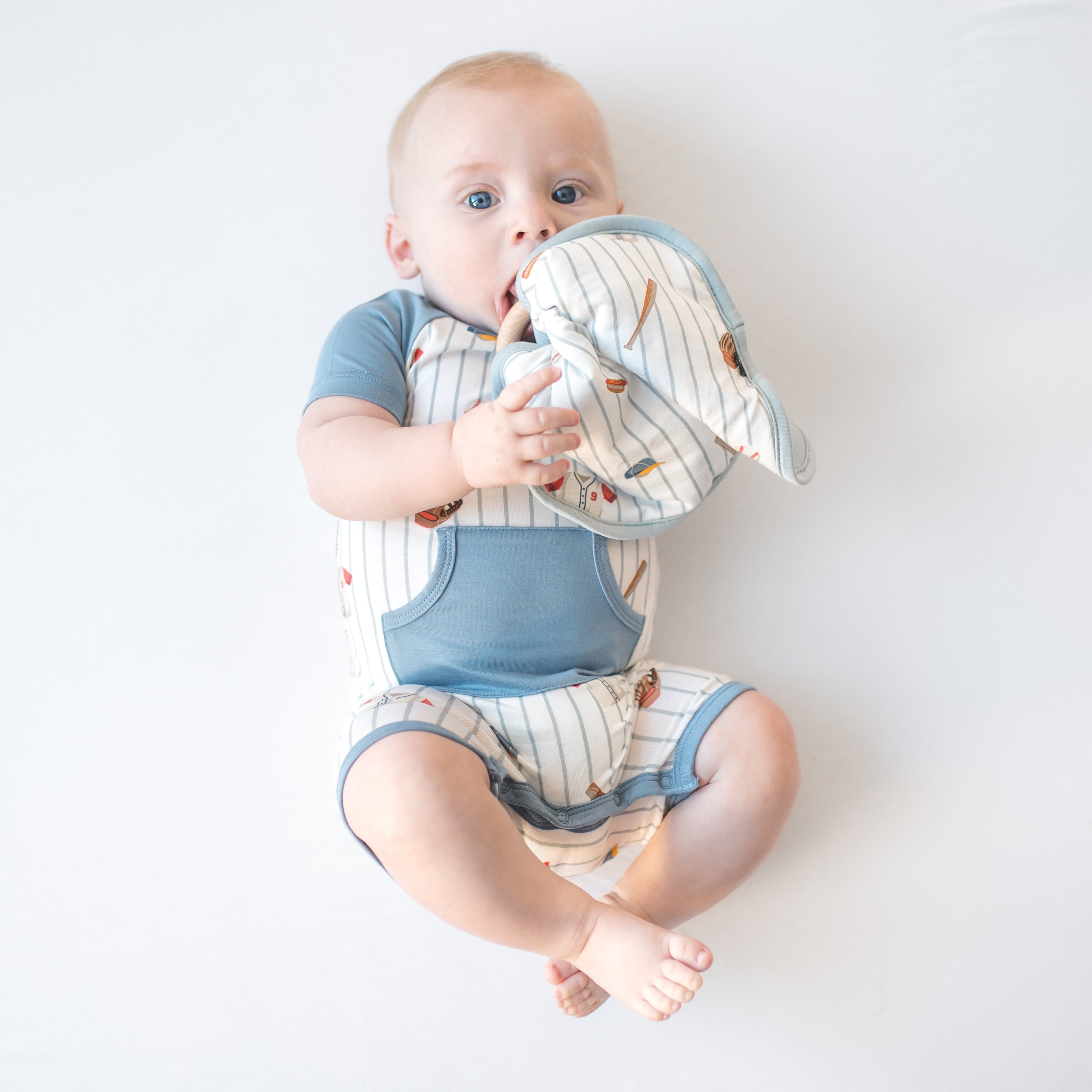 Young infant laying on a white surface holding the Lovey in Home Run with Removable Wooden Teething Ring wearing a matching shortall