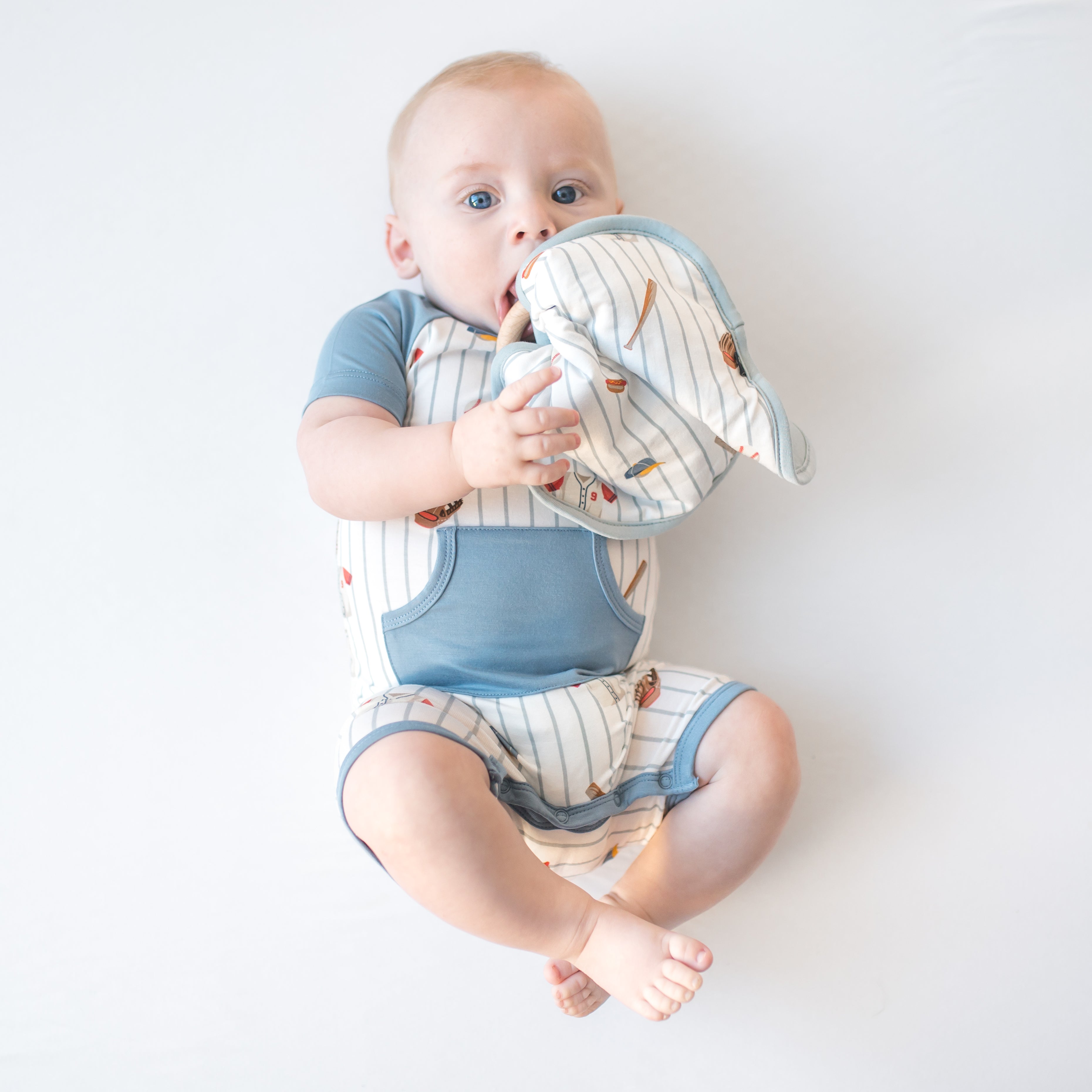 Young infant laying on a white surface holding the Lovey in Home Run with Removable Wooden Teething Ring wearing a matching shortall