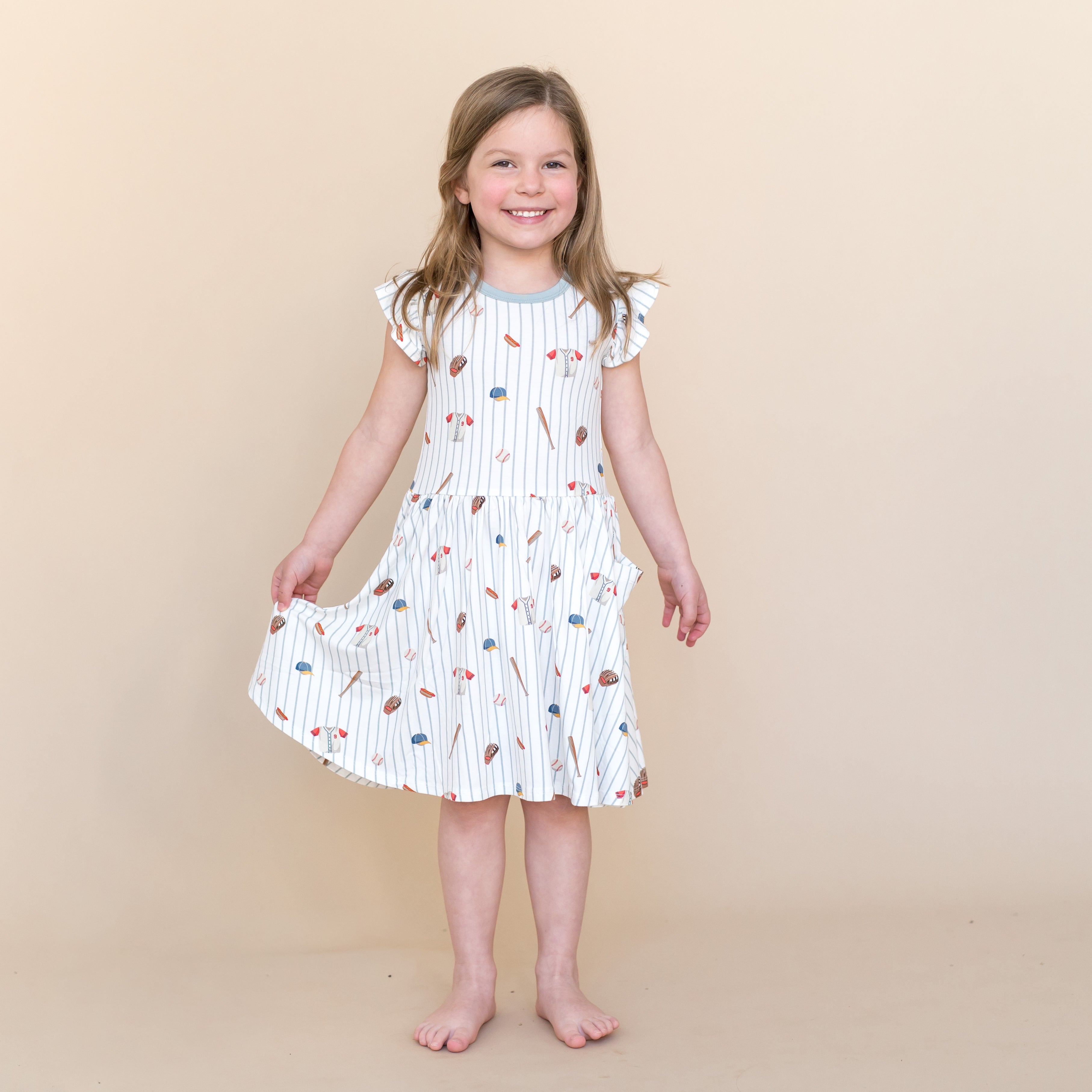 Young girl standing wearing the Pocket Dress in Home Run holding the skirt of the dress in one hand