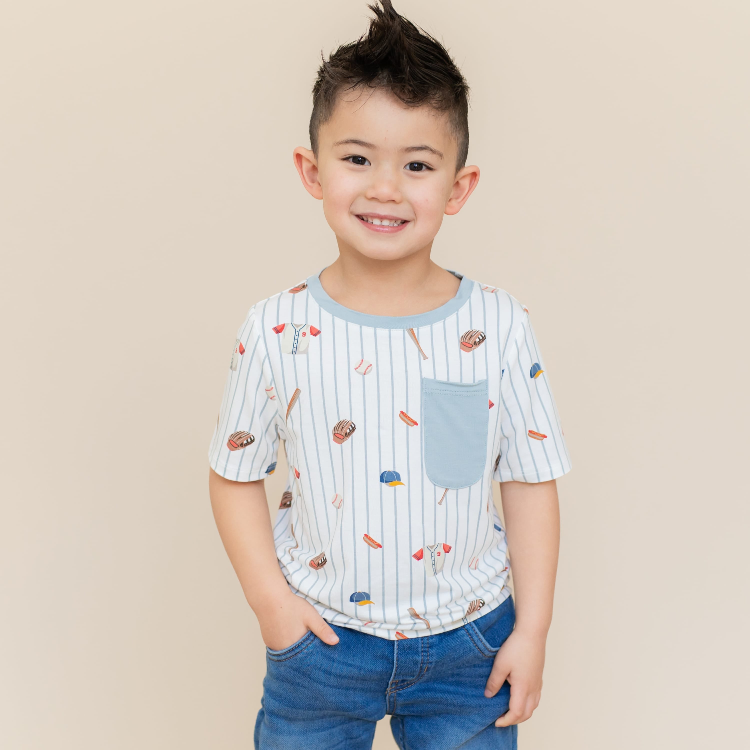 Young boy standing in front of a light beige background wearing the Toddler Crew Neck Tee in Home Run with medium wash jeans