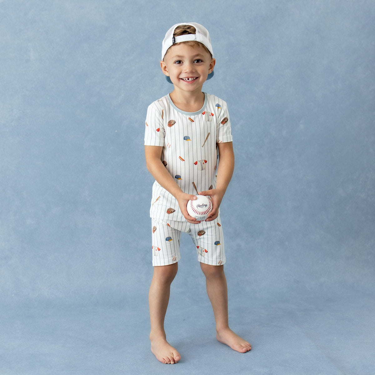 Young boy wearing the Short Sleeve Pajamas in Home Run holding a baseball wearing a backwards baseball cap