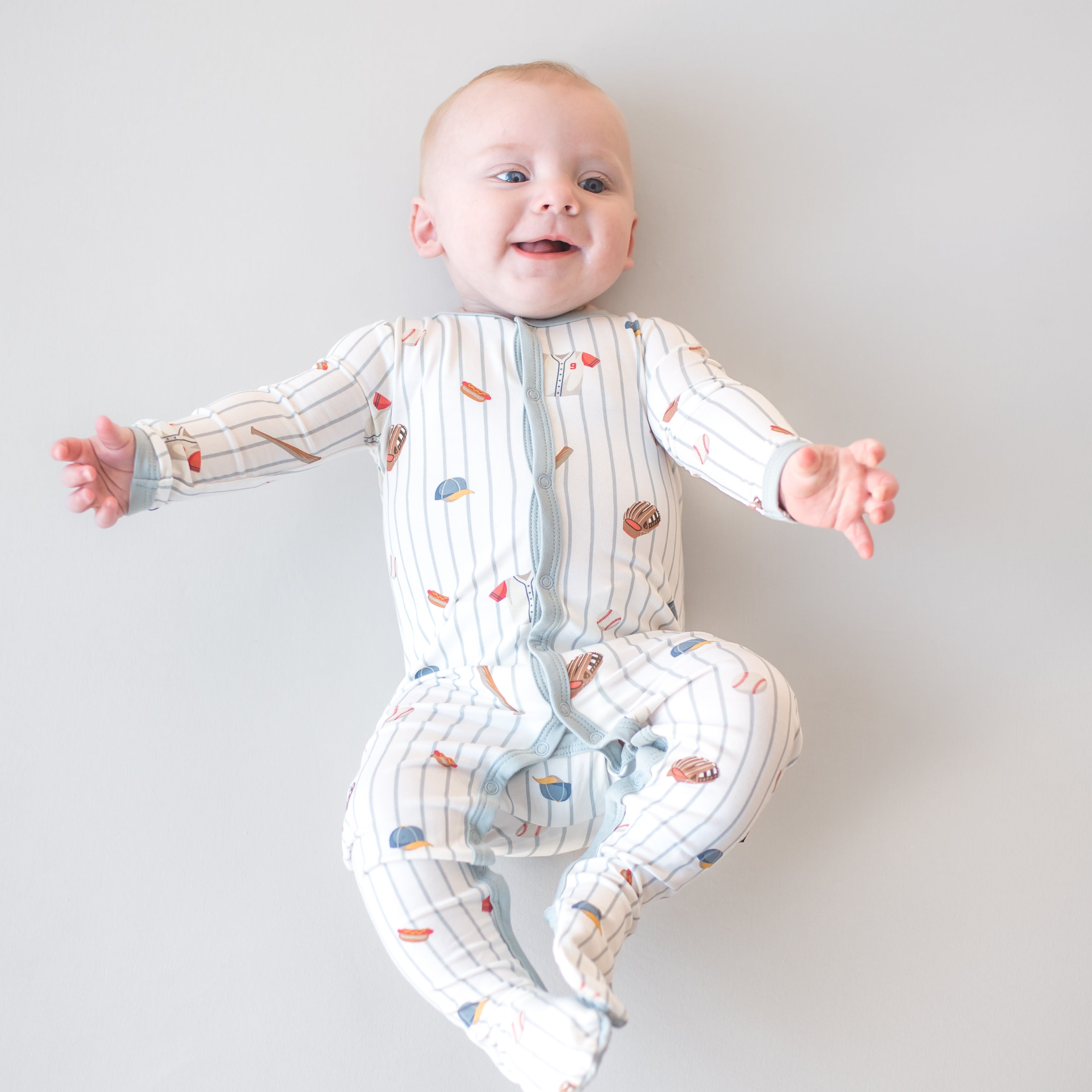 Smiling infant laying on a light surface wearing the Footie in Home Run