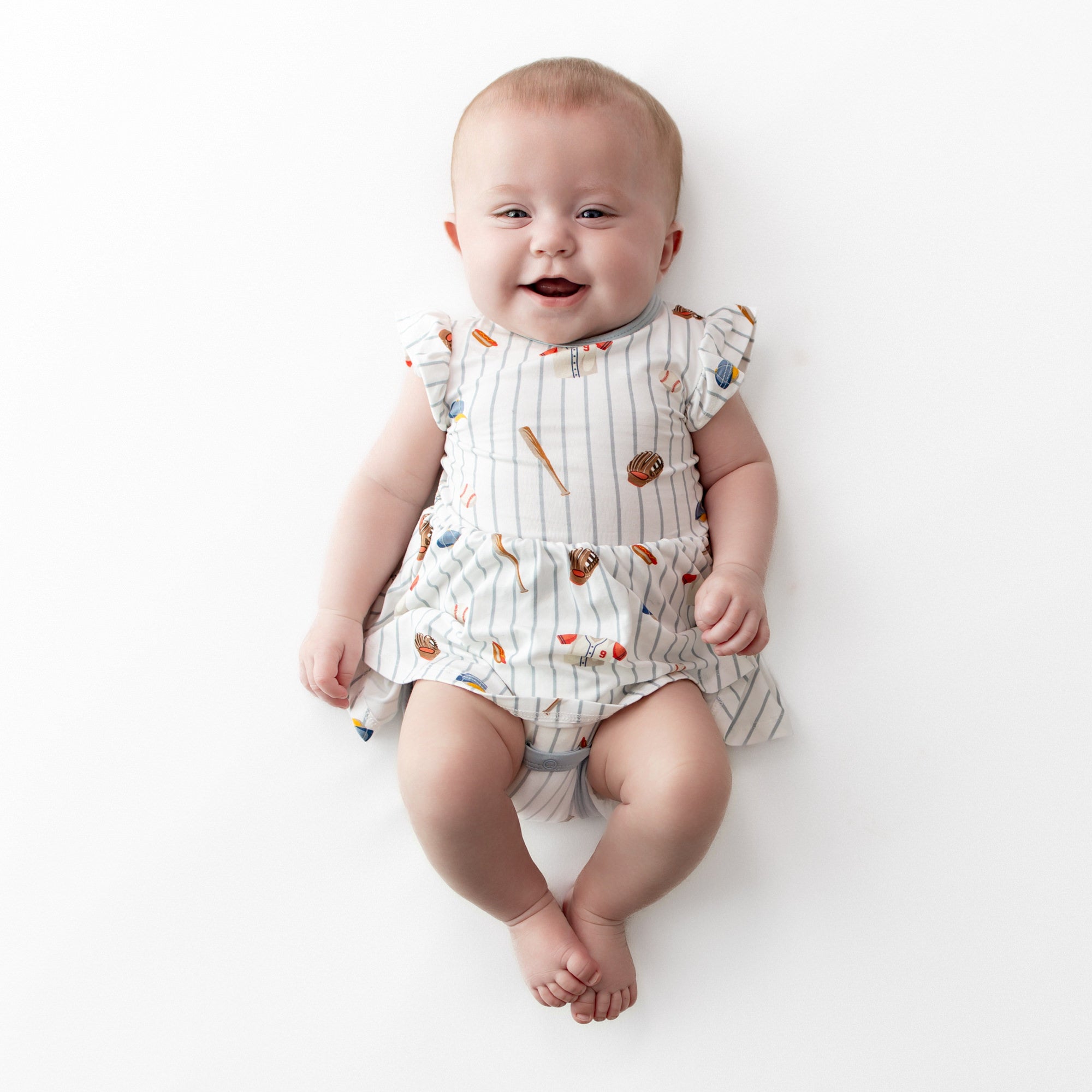 Smiling infant wearing the Twirl Bodysuit Dress in Home Run laying on a white surface