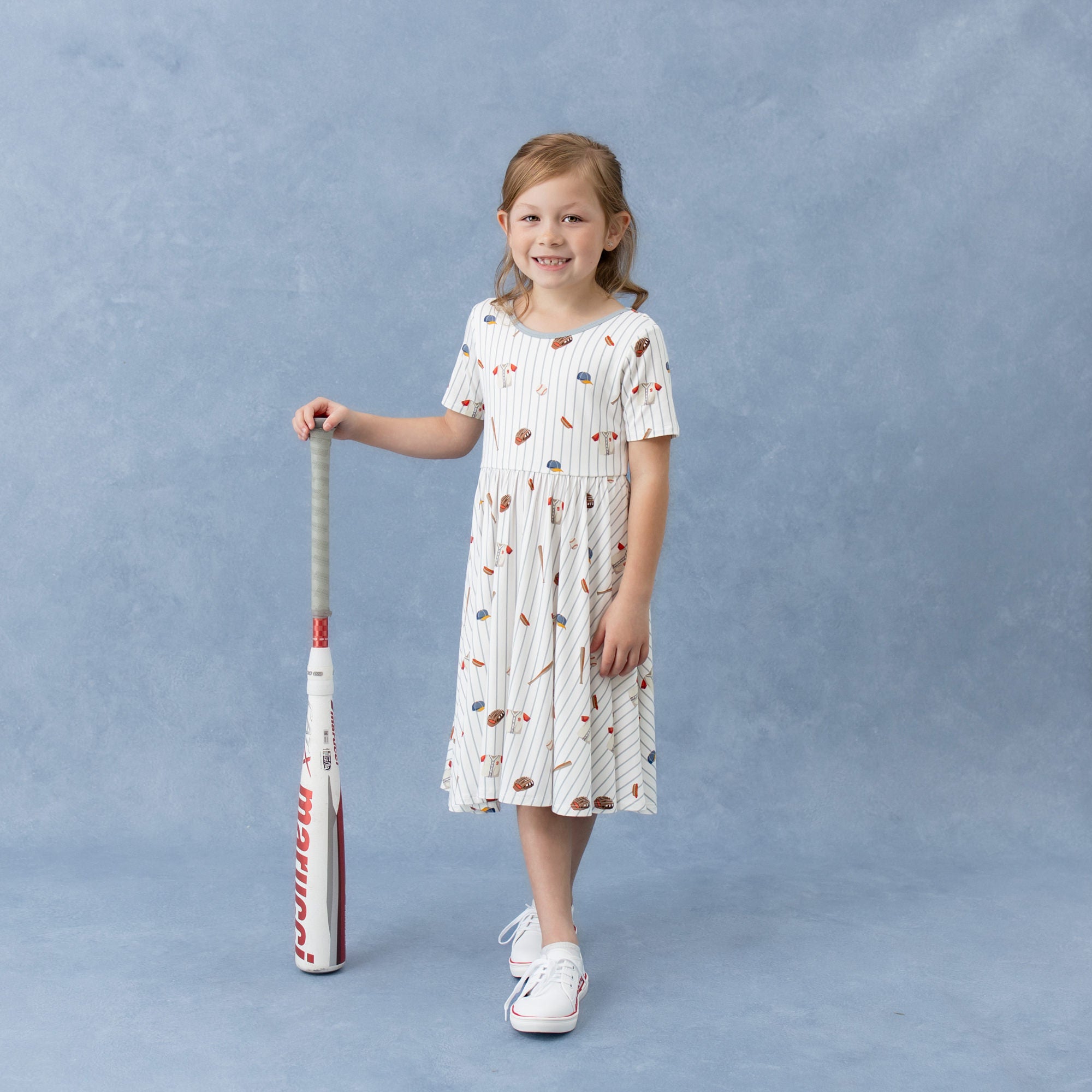 Young girl wearing the Twirl Dress in Home Run standing beside a baseball bat