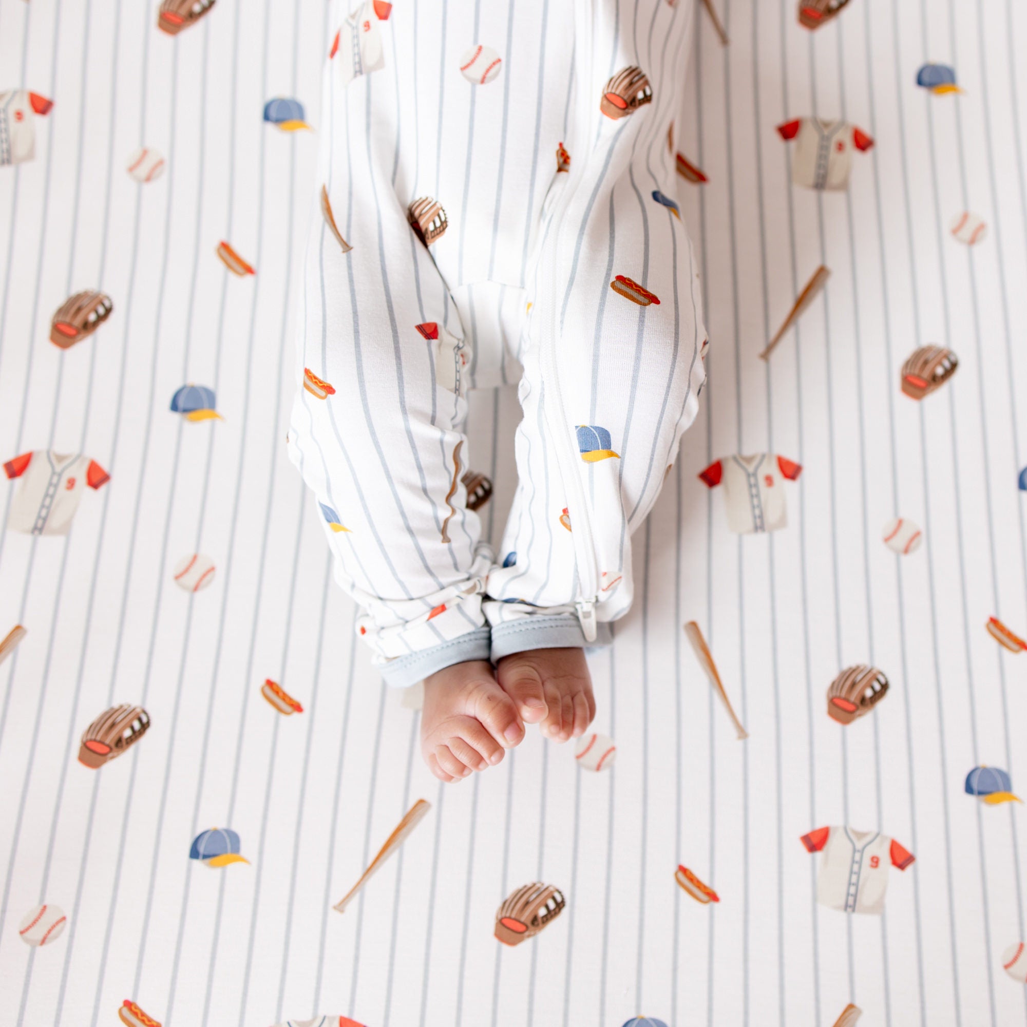 The zipper closure on the leg of the Zippered Romper in Home Run shown on an infant model laying on a Crib Sheet in Home Run
