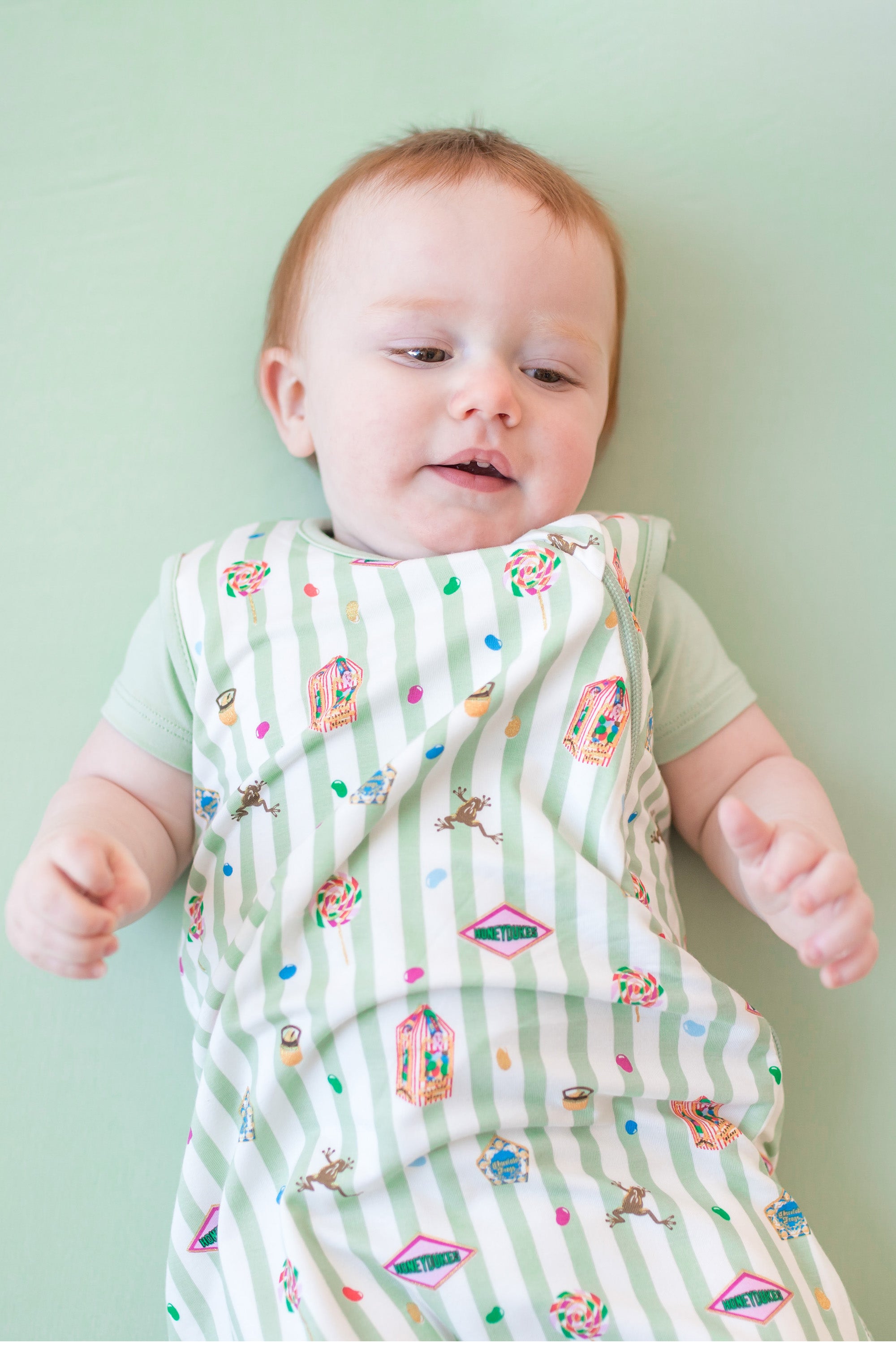 Baby wearing a green and white striped sleep bag with colorful Harry Potter patterns on a green background