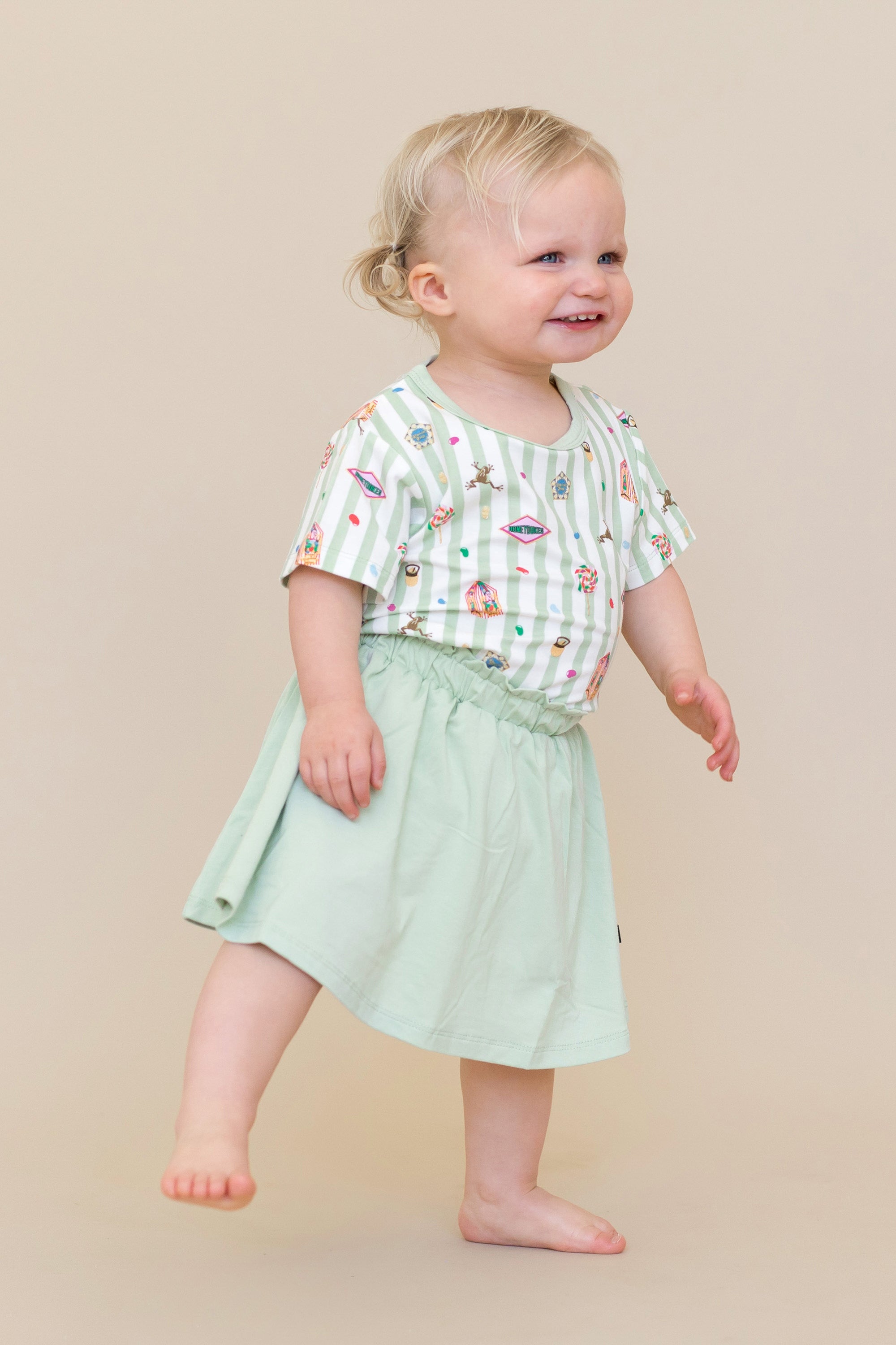Child wearing a green and white striped Toddler Basic Tee with colorful Harry Potter patterns with a green skort on a beige background