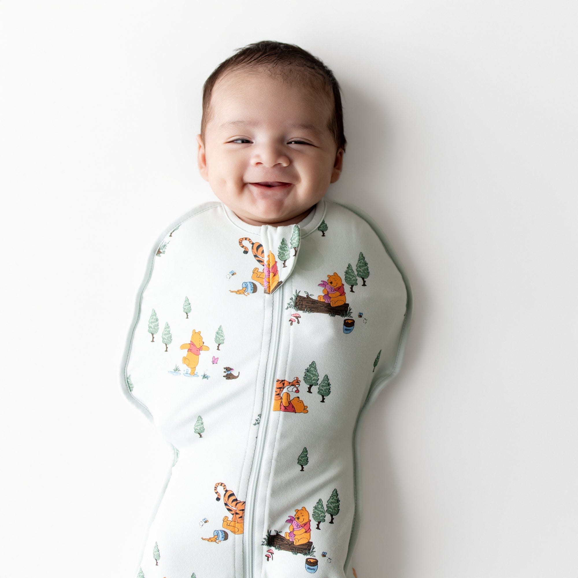 Close up of a smiling infant wearing the Swaddle Bag in Winnie the Pooh Hundred Acre Wood 0.5