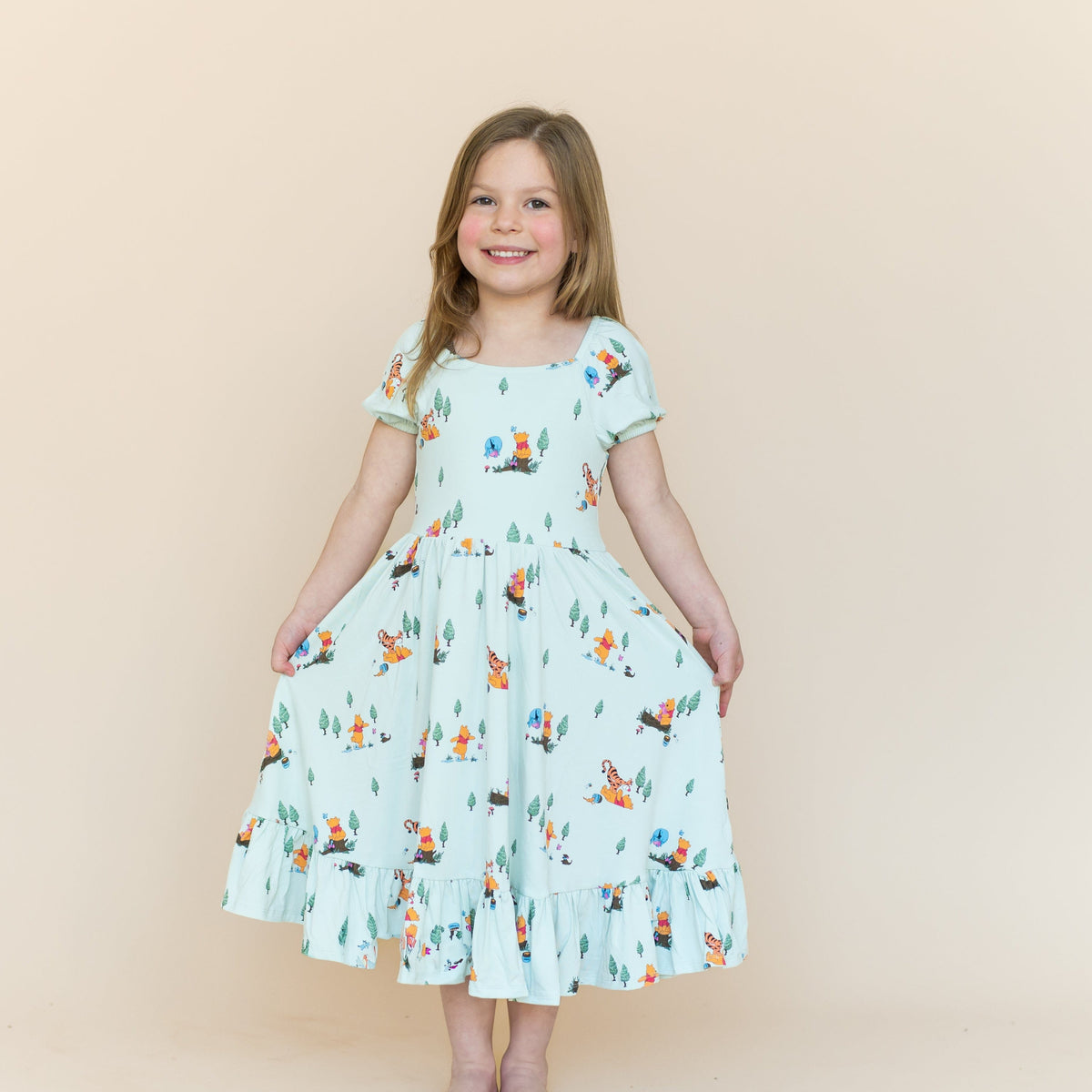 Young smiling girl wearing the Puff Sleeve Twirl Dress in Winnie the Pooh Hundred Acre Wood holding the skirt of the dress out
