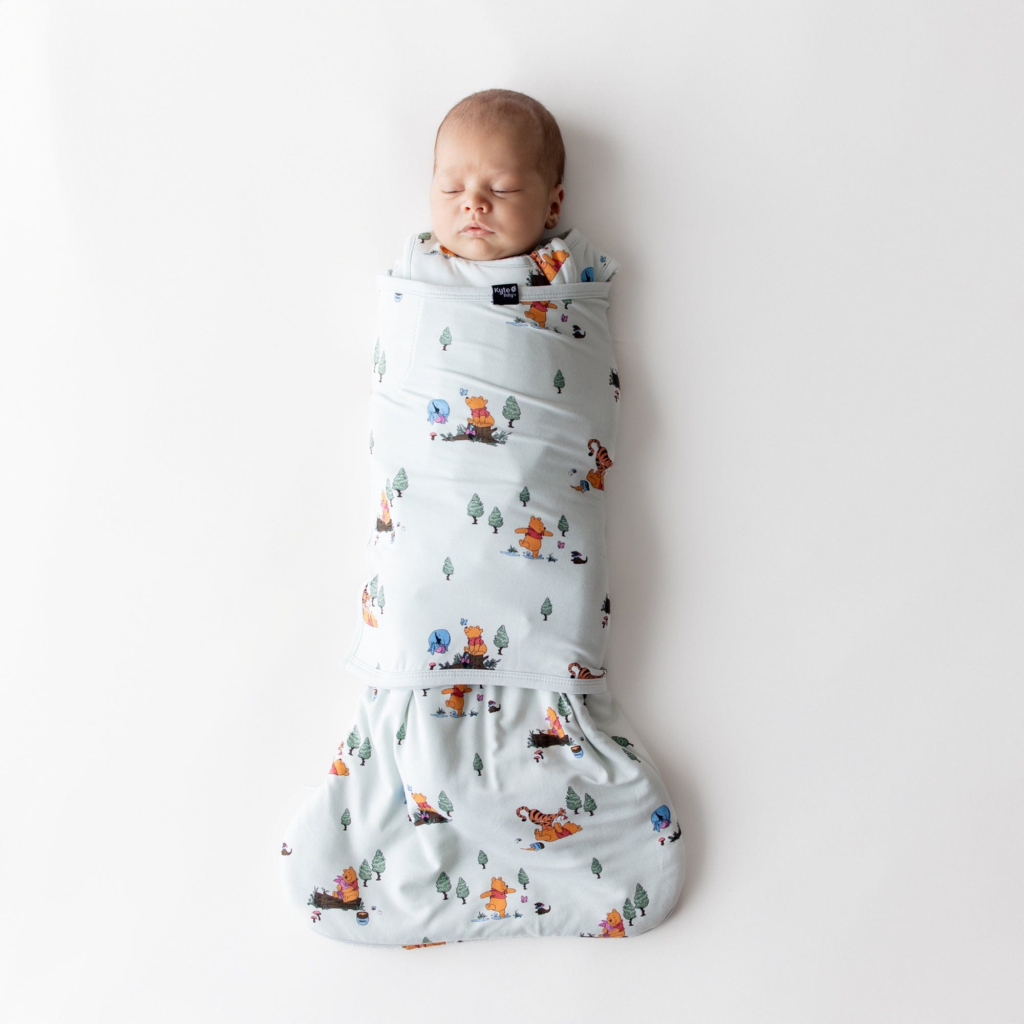 Sleeping newborn swaddled in the Sleep Bag Swaddler in Winnie the Pooh Hundred Acre Wood 1.0 laying on a white surface