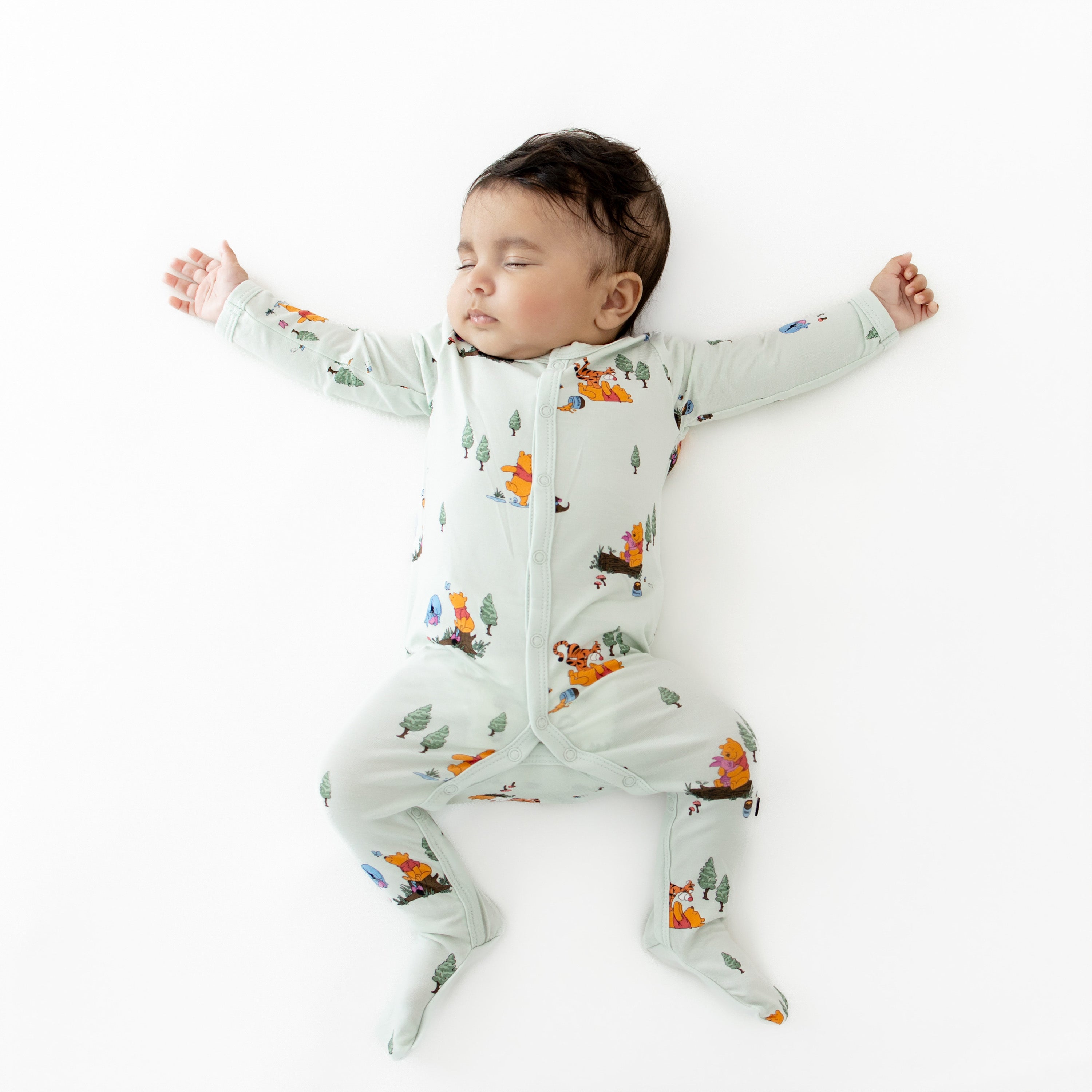 Sleeping infant stretched out wearing the Footie in Winnie the Pooh Hundred Acre Wood on a white surface