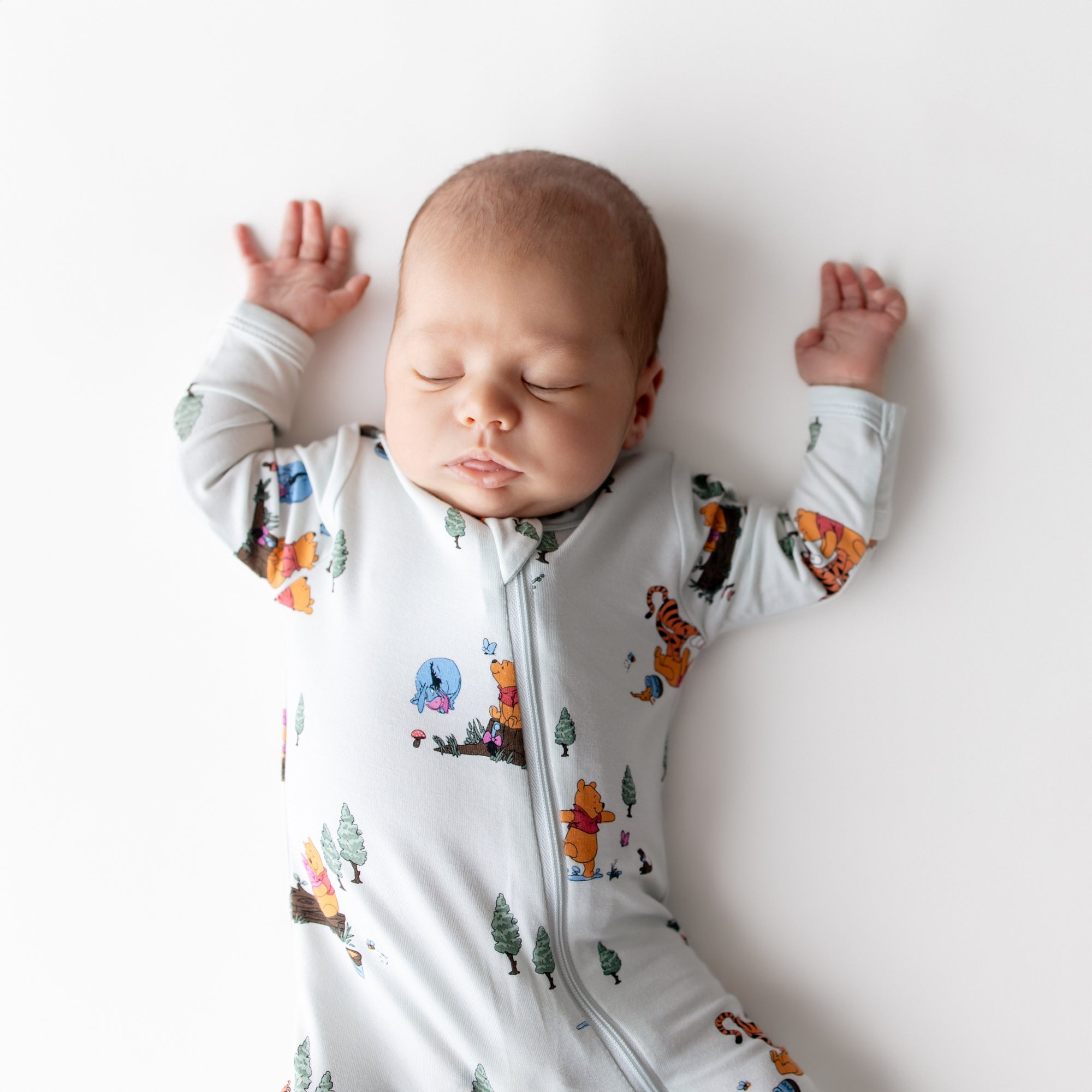 Close up of sleeping newborn with their arms up wearing the Zippered Footie in Winnie the Pooh Hundred Acre Wood