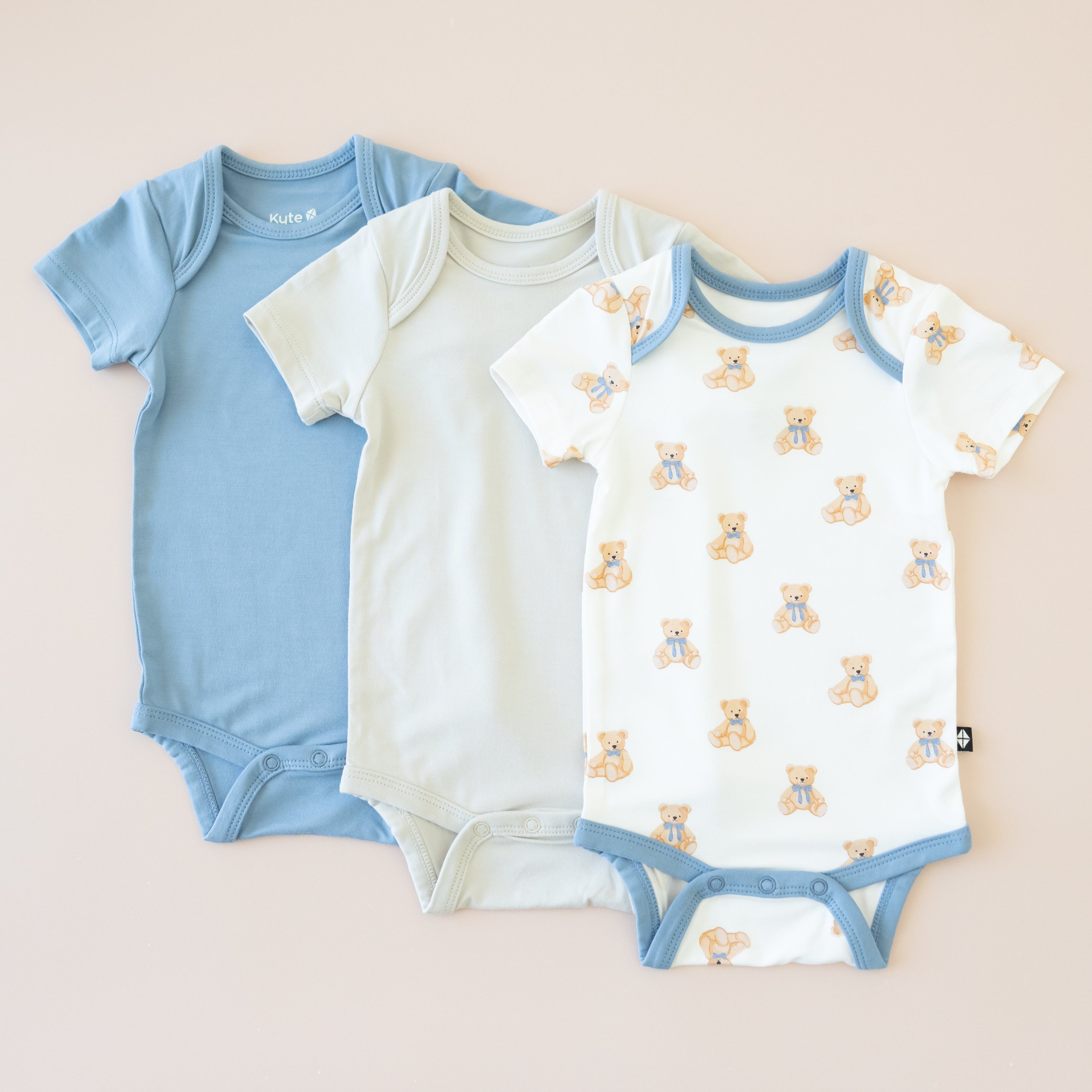 Three baby bodysuits in blue, off-white, and patterned with teddy bears on a beige background.