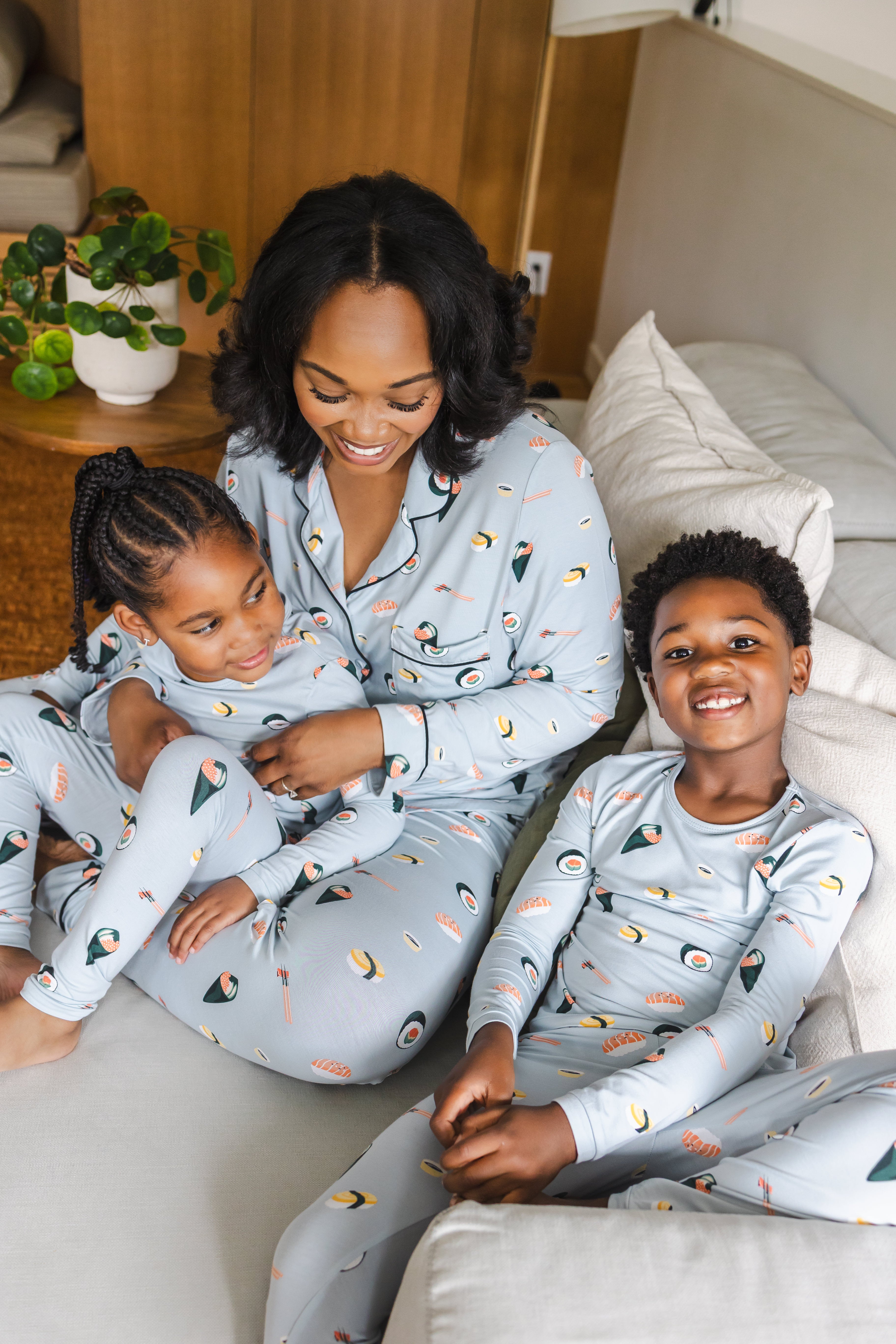 A woman and two children wearing matching light blue pajamas with a colorful sushi print, sitting together on a light-colored sofa in a cozy living room setting.