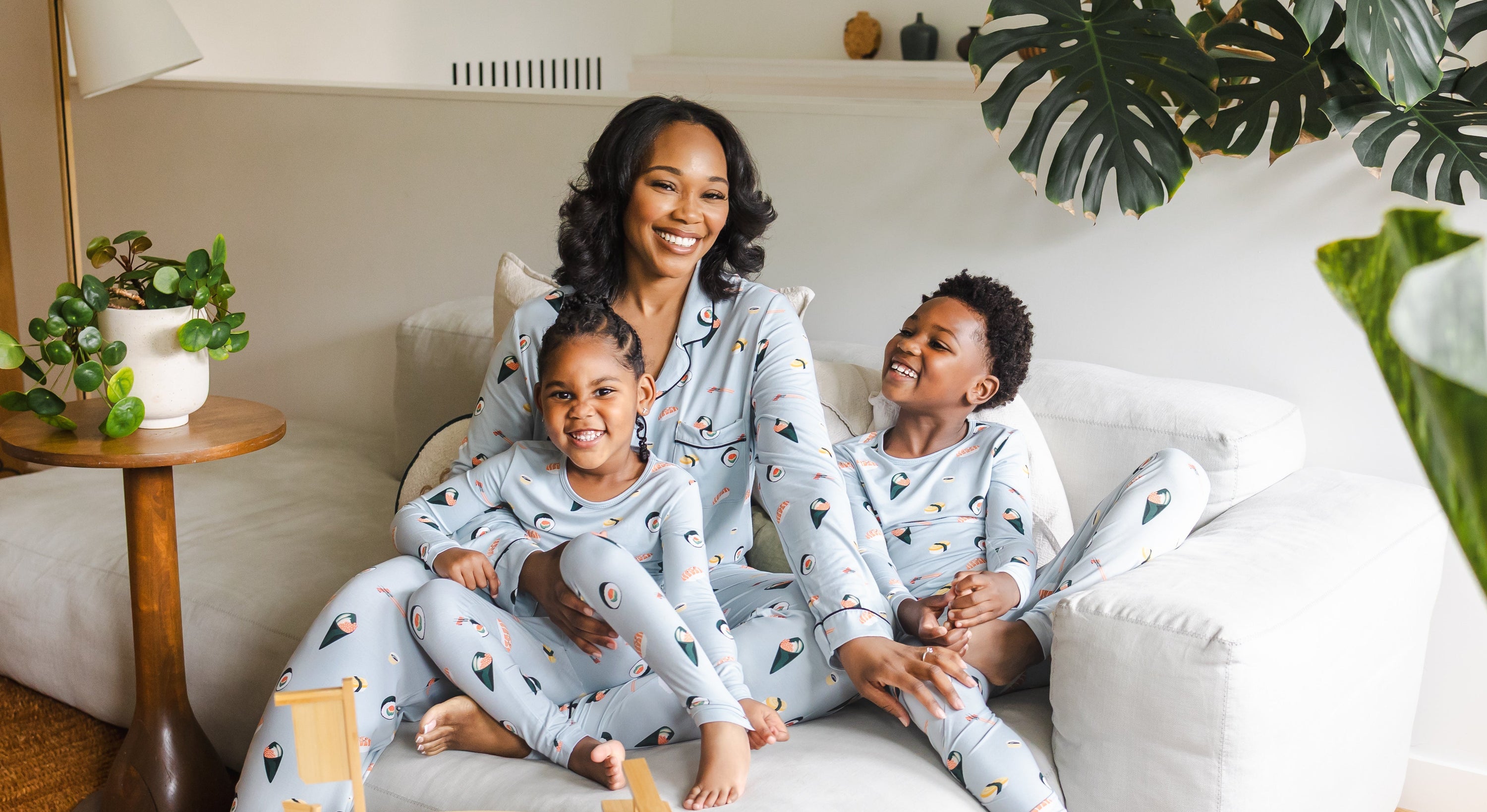 A family of three wearing matching light blue pajamas with a colorful sushi print sits together on a white sofa in a bright living room. A small wooden table in the foreground holds a sushi boat prop and small dishes.