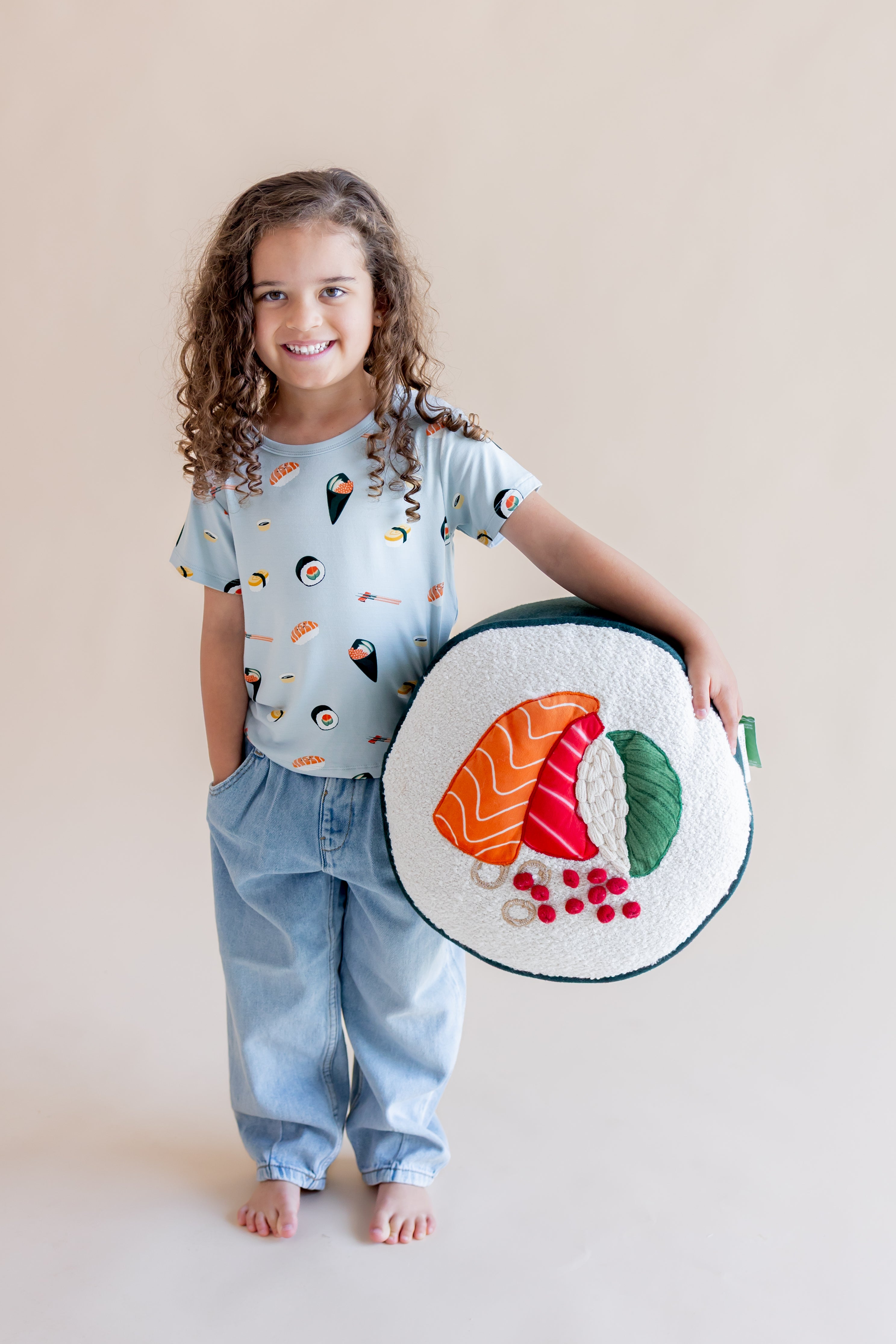 Child smiling in ice sushi tee, jeans, hand in a pocket with the other hand holding a sushi pillow
