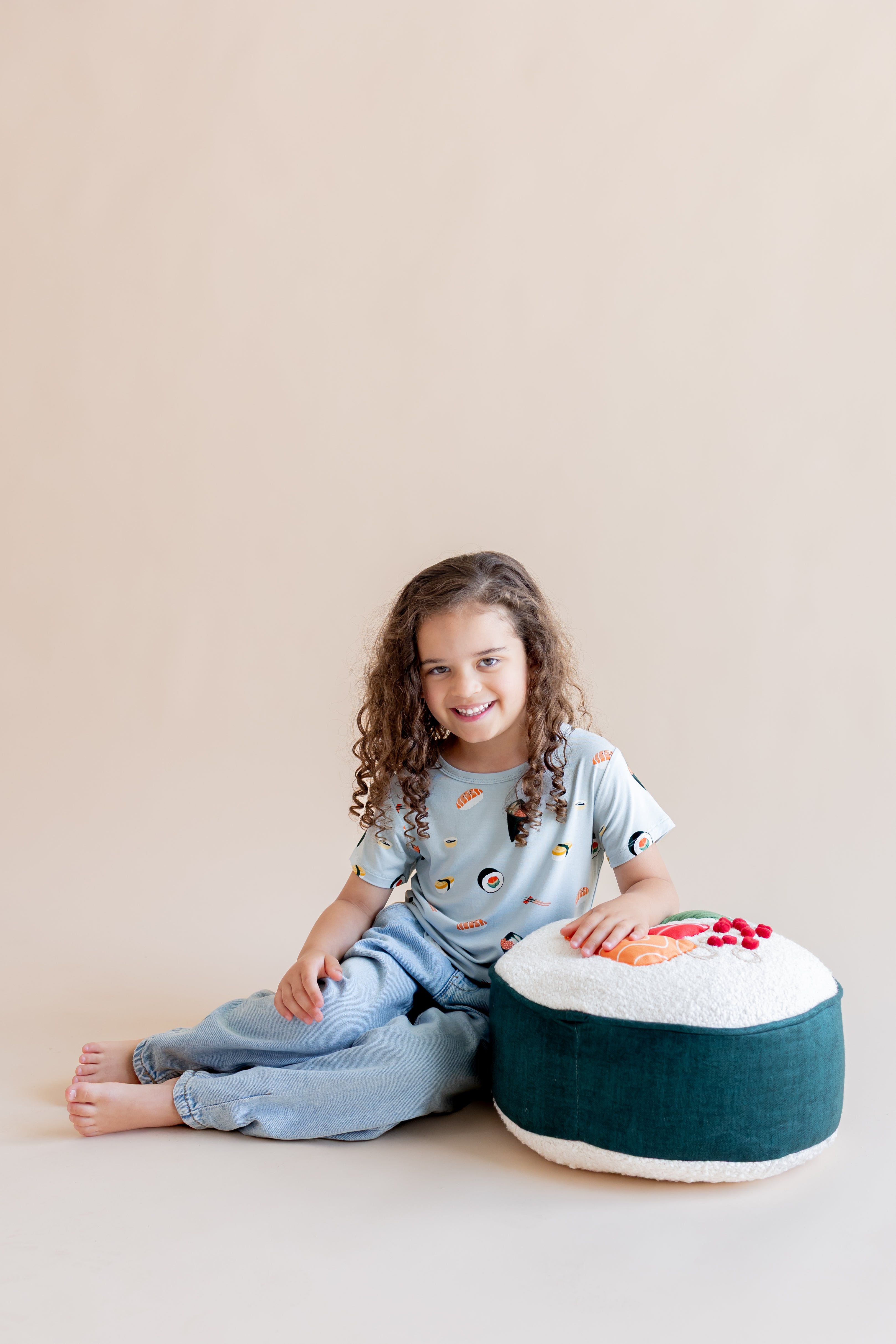 Child smiling sitting on their side leaning on a sushi pillow in ice sushi tee and pants